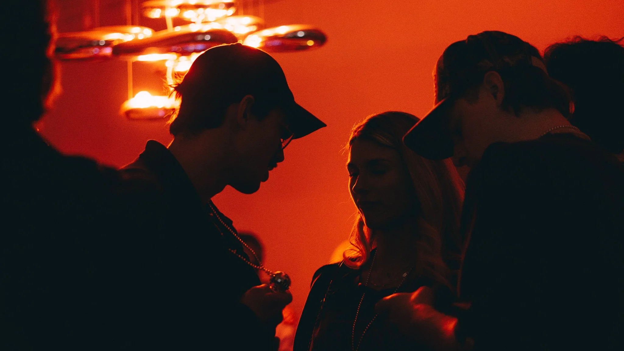 Silhouettes of four people in a dimly lit room with orange lighting, engaging in conversation, with a chandelier or ceiling lights in the background.