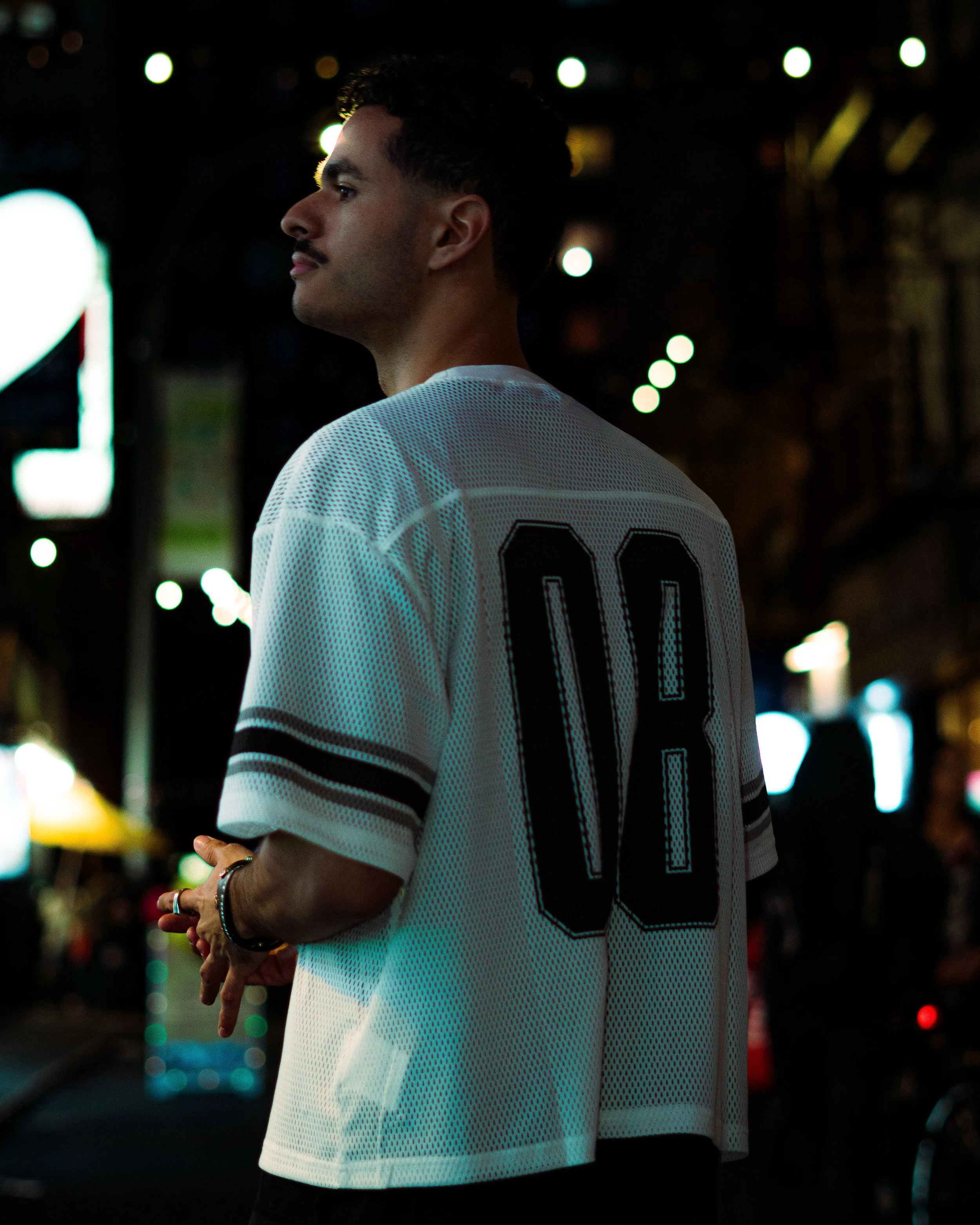 A young man with short dark hair and a mustache stands on a city street at night with blurred lights behind him, wearing a white sports jersey with the number 00 on the back.
