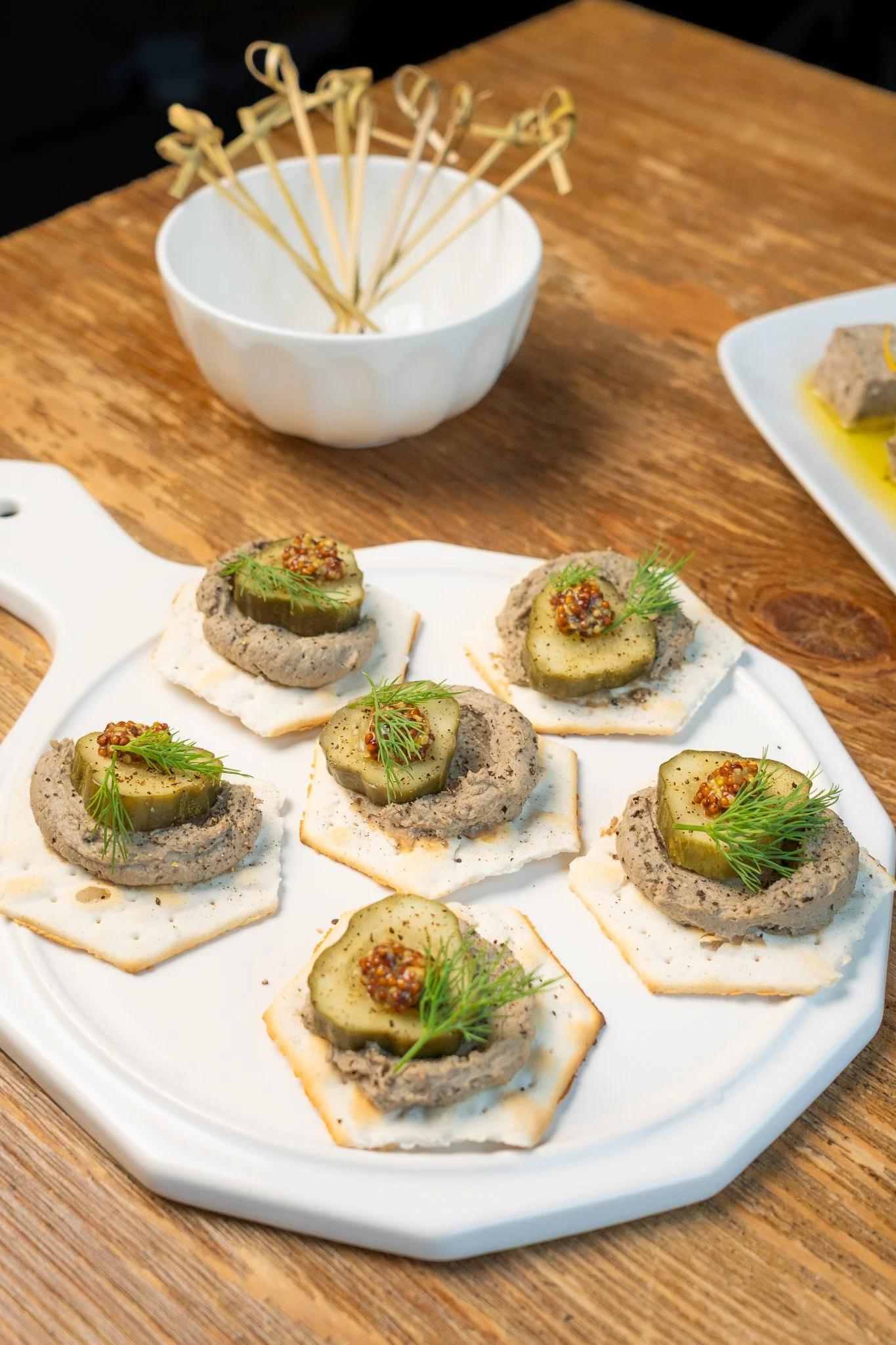 Appetizer bites on a white plate, topped with pickles, mustard seeds, and herbs, arranged on a wooden table.