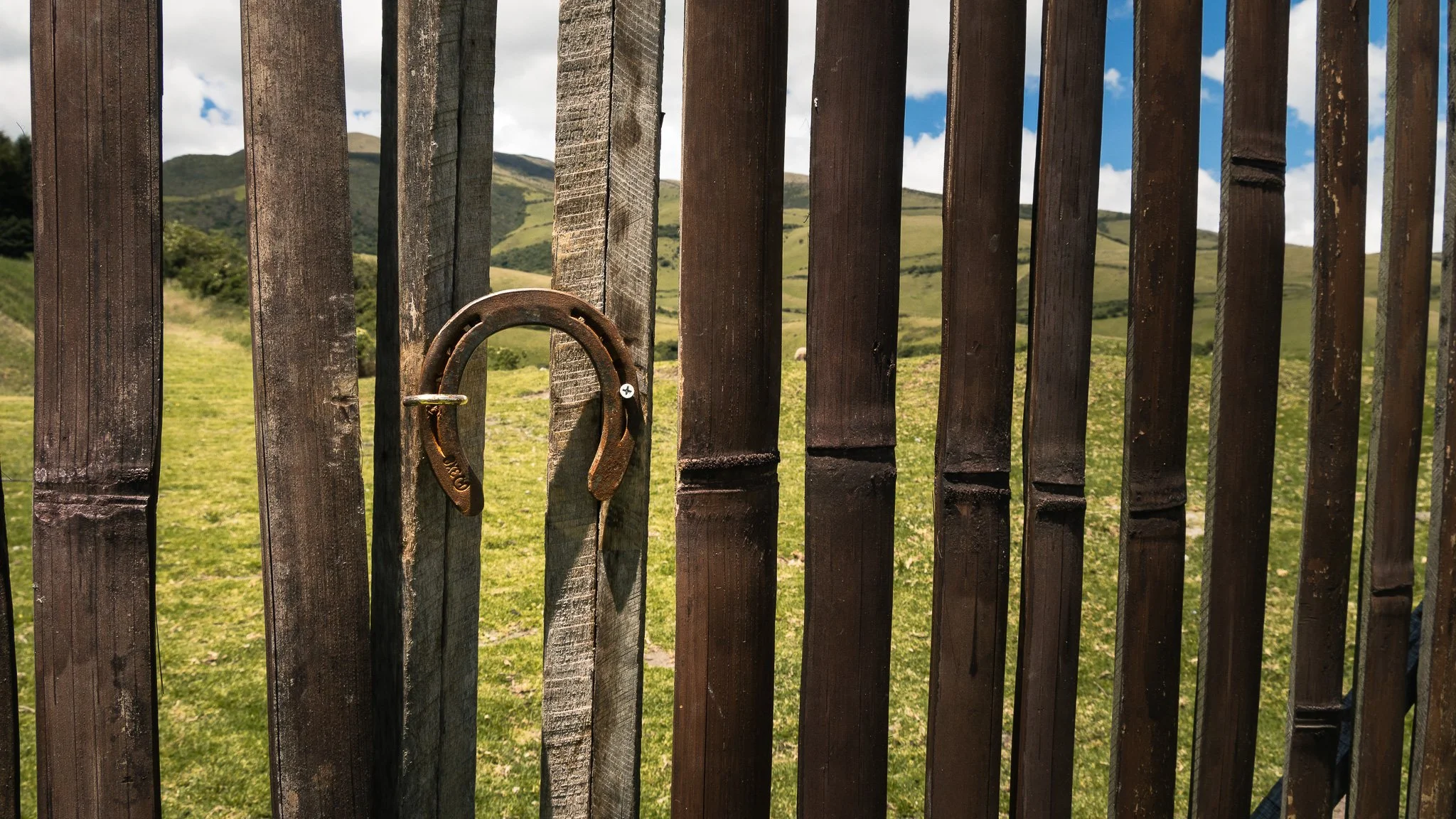 A wooden gate with a rusty horseshoe hanging on one of the vertical slats, overlooking green rolling hills and mountains under a partly cloudy sky.
