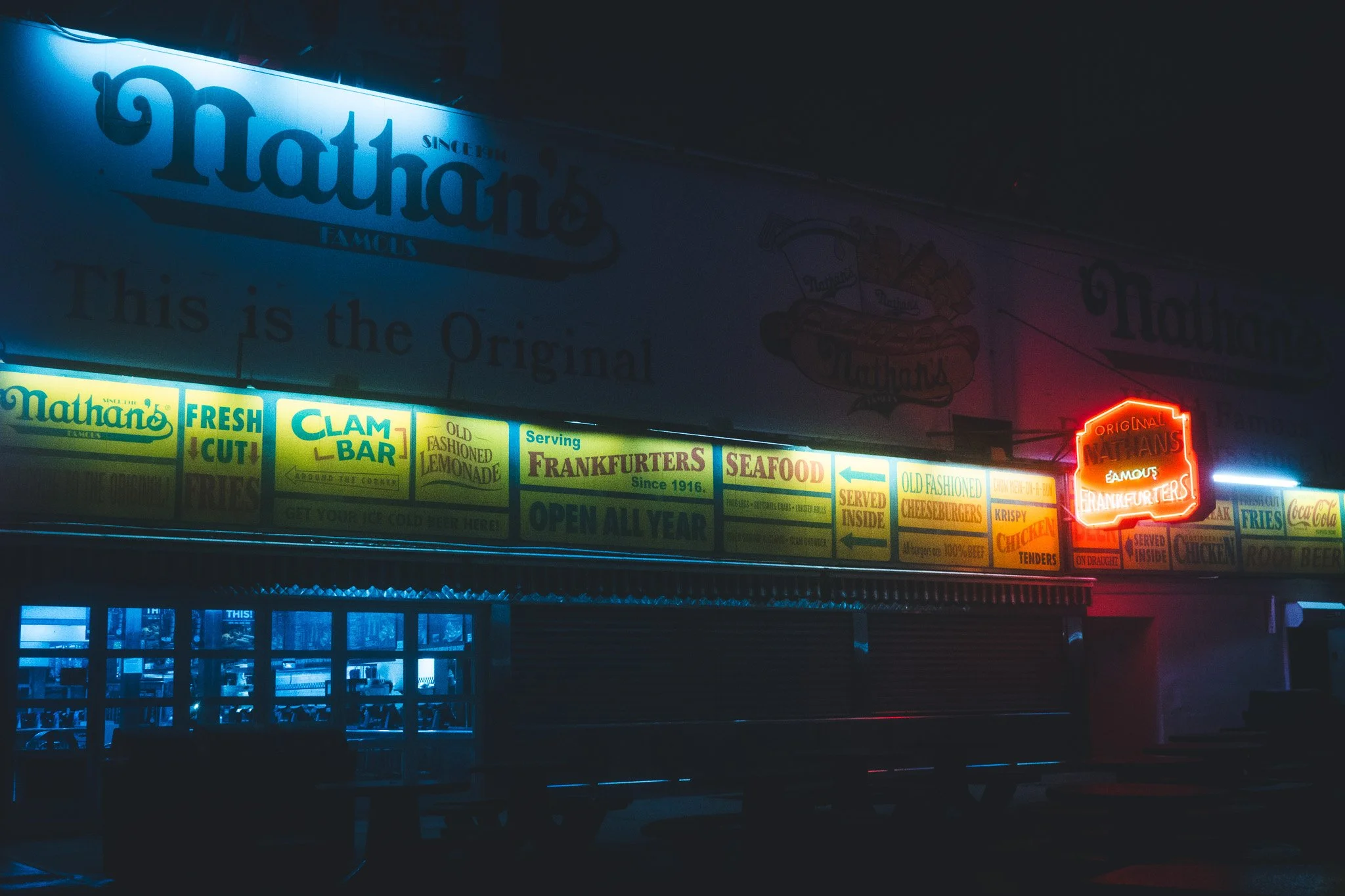 An illuminated sign for Nathan's Famous, a seafood restaurant, at night. The sign features blue, yellow, and red neon lights and displays various food items and slogans.