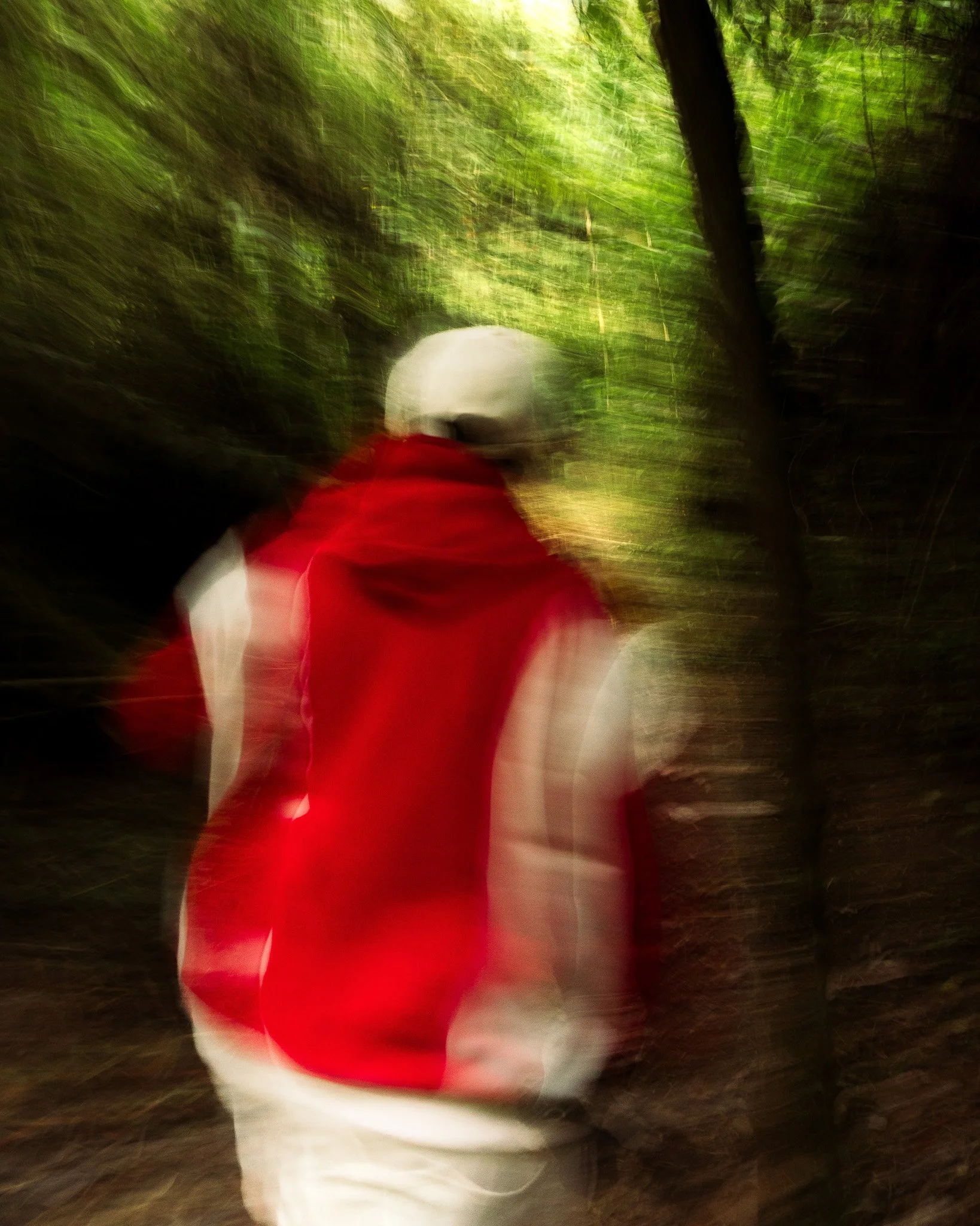 A motion-blurred image of a person wearing a red and white outfit, moving through a forested trail with lush green trees.