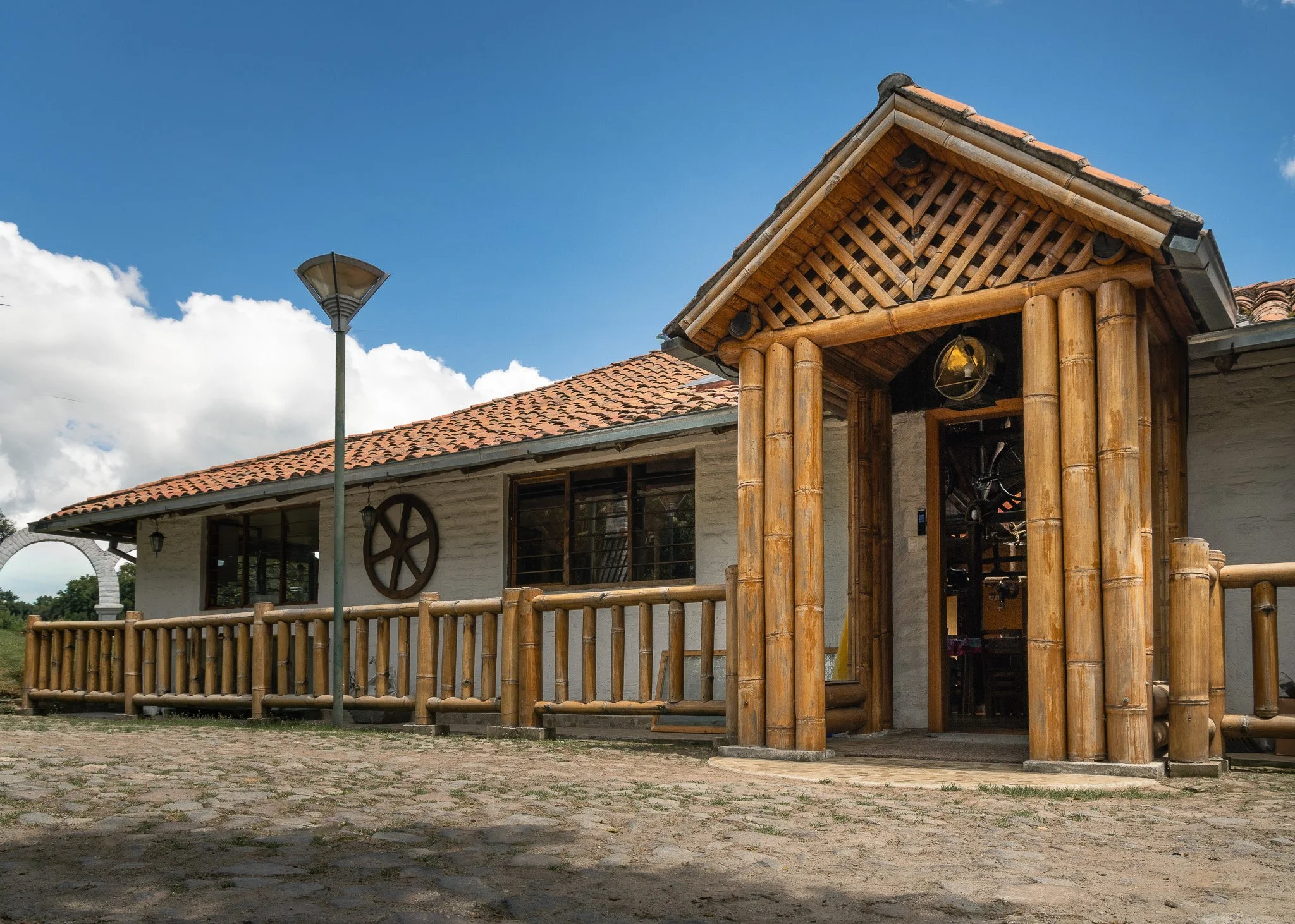 A house with a white exterior, a tiled roof, and a wooden porch with bamboo poles. There is a street lamp and a wheel decoration on the house’s wall. The sky is blue with some clouds.