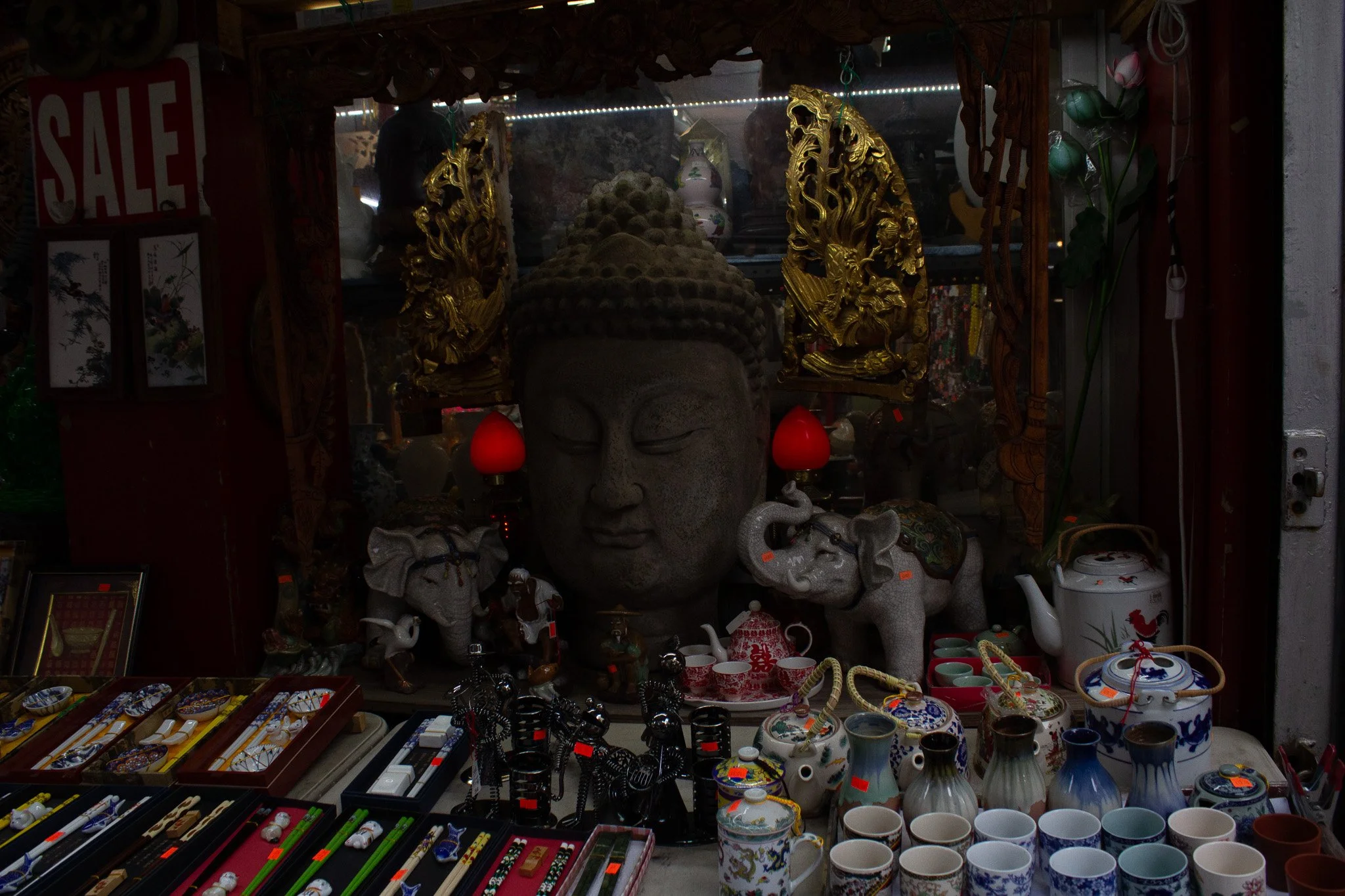 A display of various items at a market stall, including a large Buddha head sculpture, decorative elephants, small teapots, cups, and jewelry with some red sale signs. Gold decorative elements and floral arrangements are also visible.