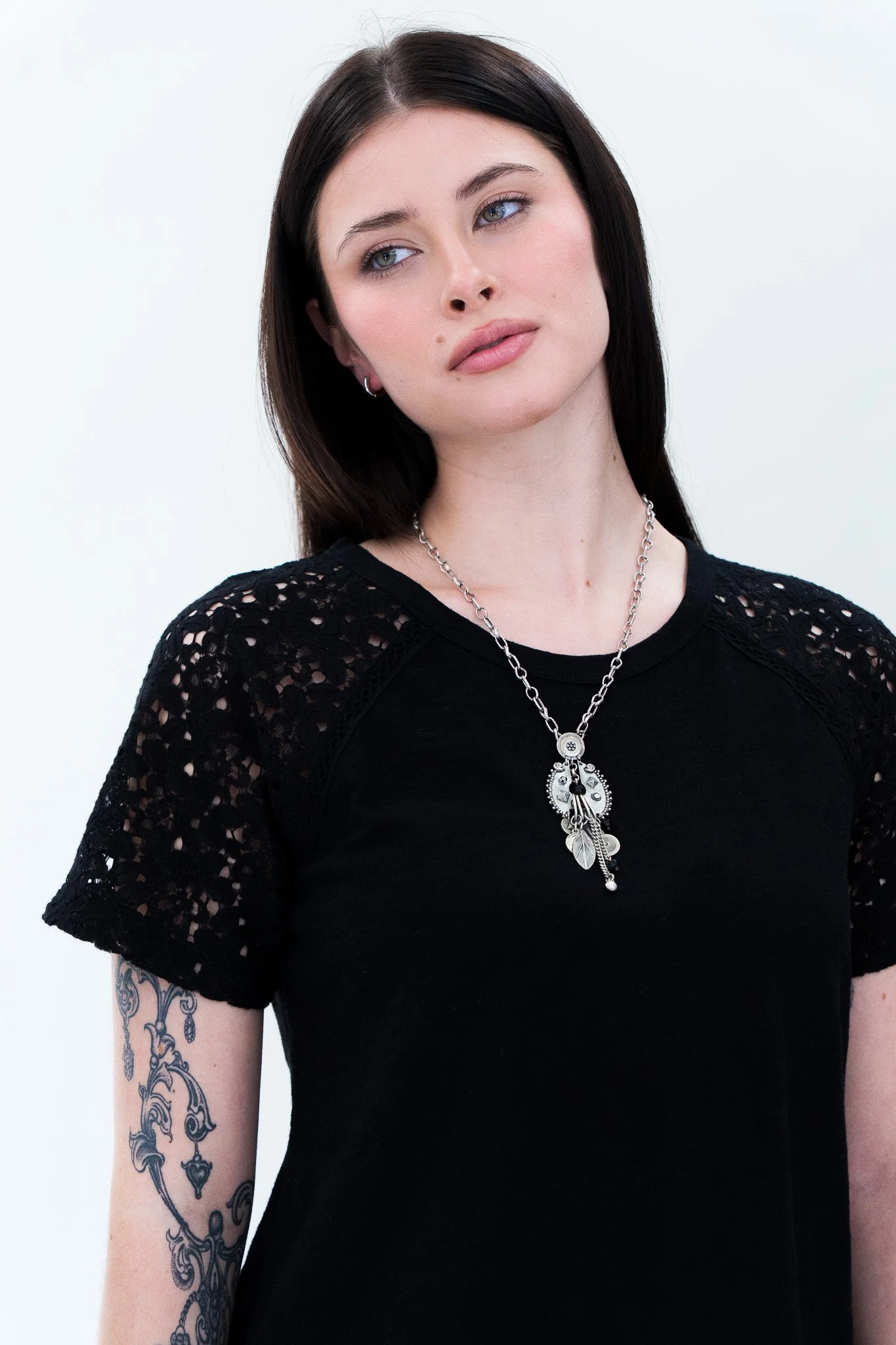 A woman with dark hair and tattoos on her arm wearing a black top with lace sleeves, a silver necklace with pendants, and small earrings, standing against a plain white background.