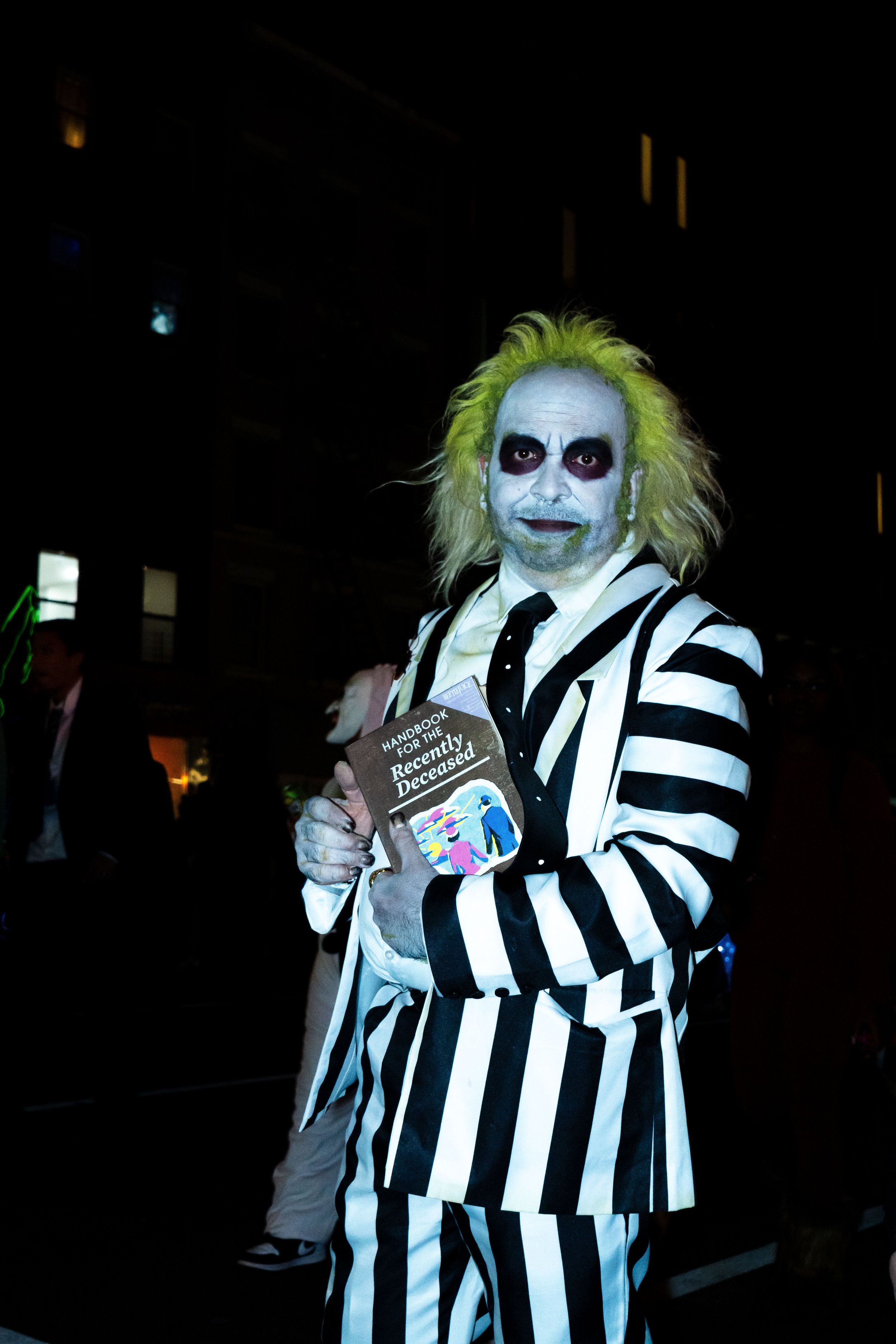 Person dressed as a clown or character with white face paint, yellow hair, and a black and white striped suit, holding a book titled 'Handbook for the Recently Deceased' at a dark event or party.