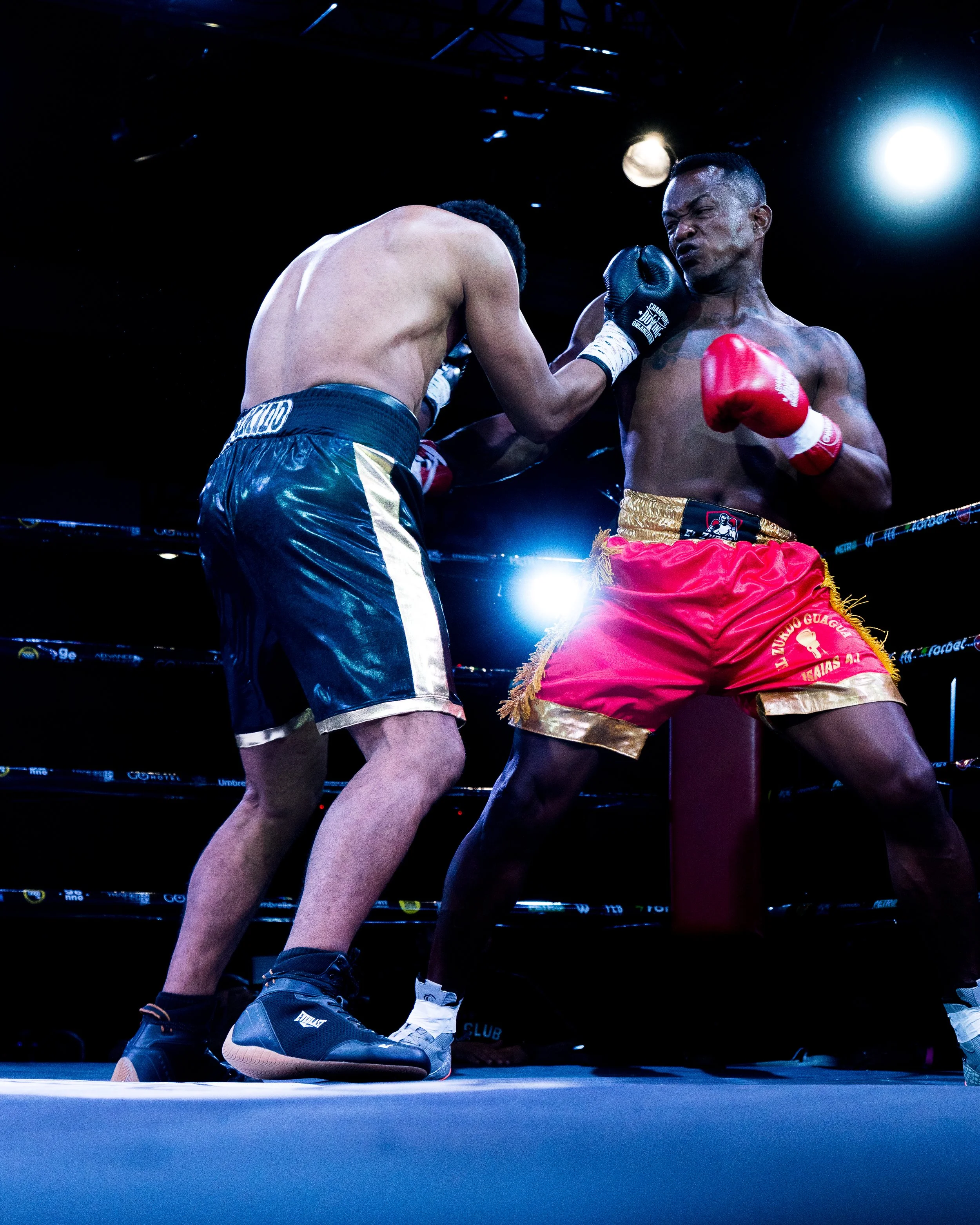 Two boxers in a ring, one throwing a punch to the other's face, with focus on their legs and gloves, under bright overhead lights.