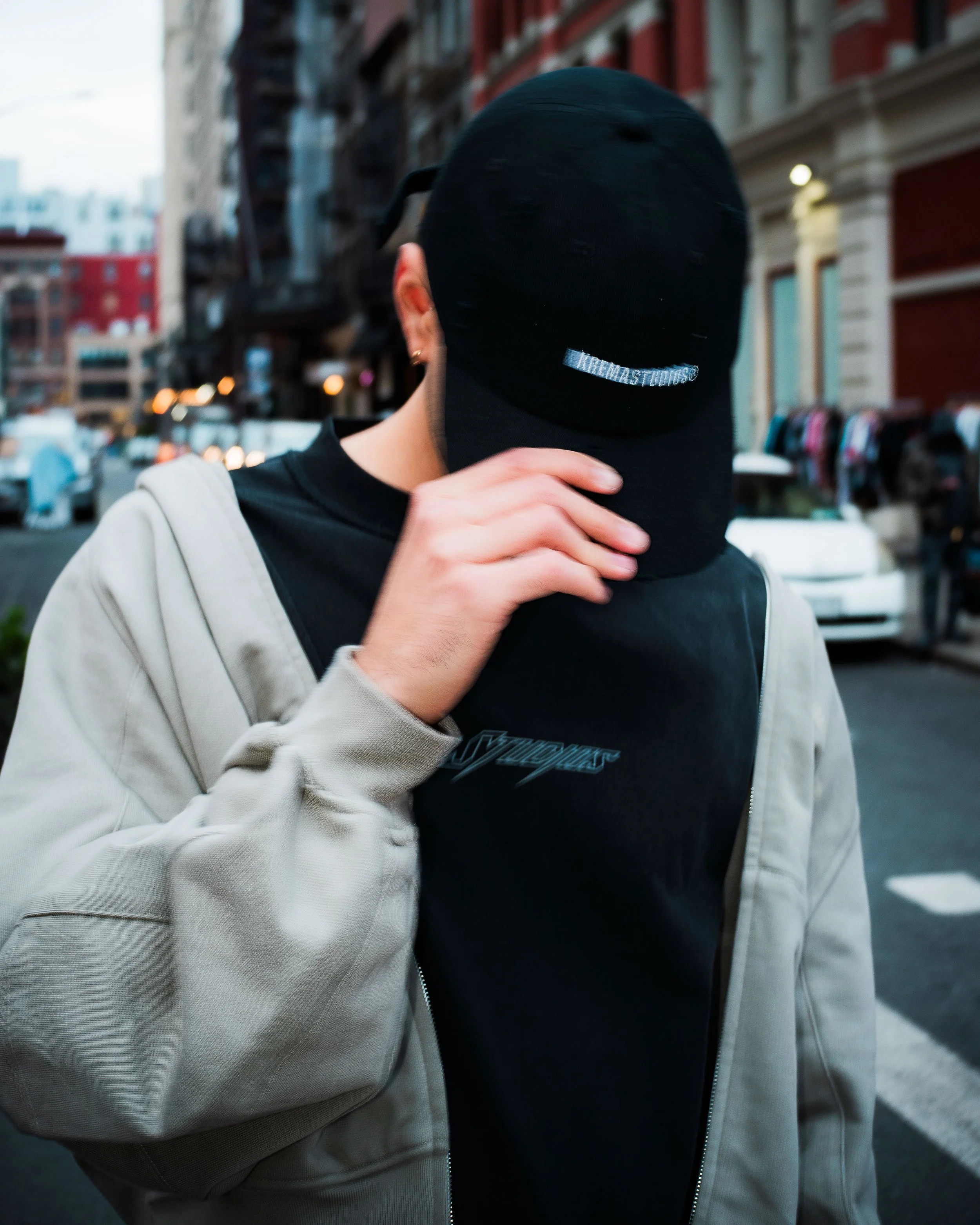 Person wearing a black cap and black t-shirt with a gray hoodie, standing on a city street with blurred buildings and cars in the background.