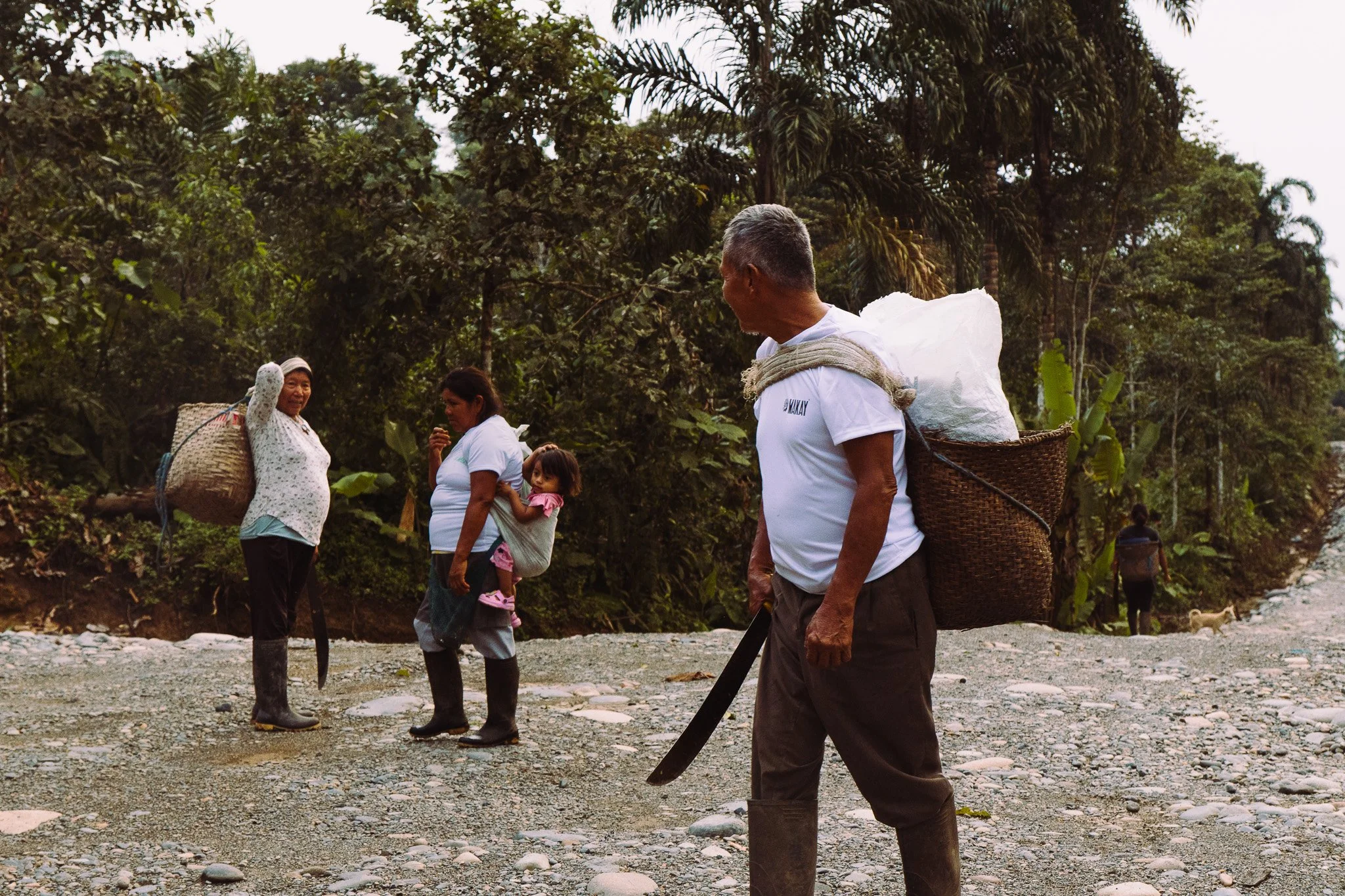 A group of people, including children and adults, standing outdoors in a lush, green forested area with trees and a rocky ground, some carrying baskets and talking.