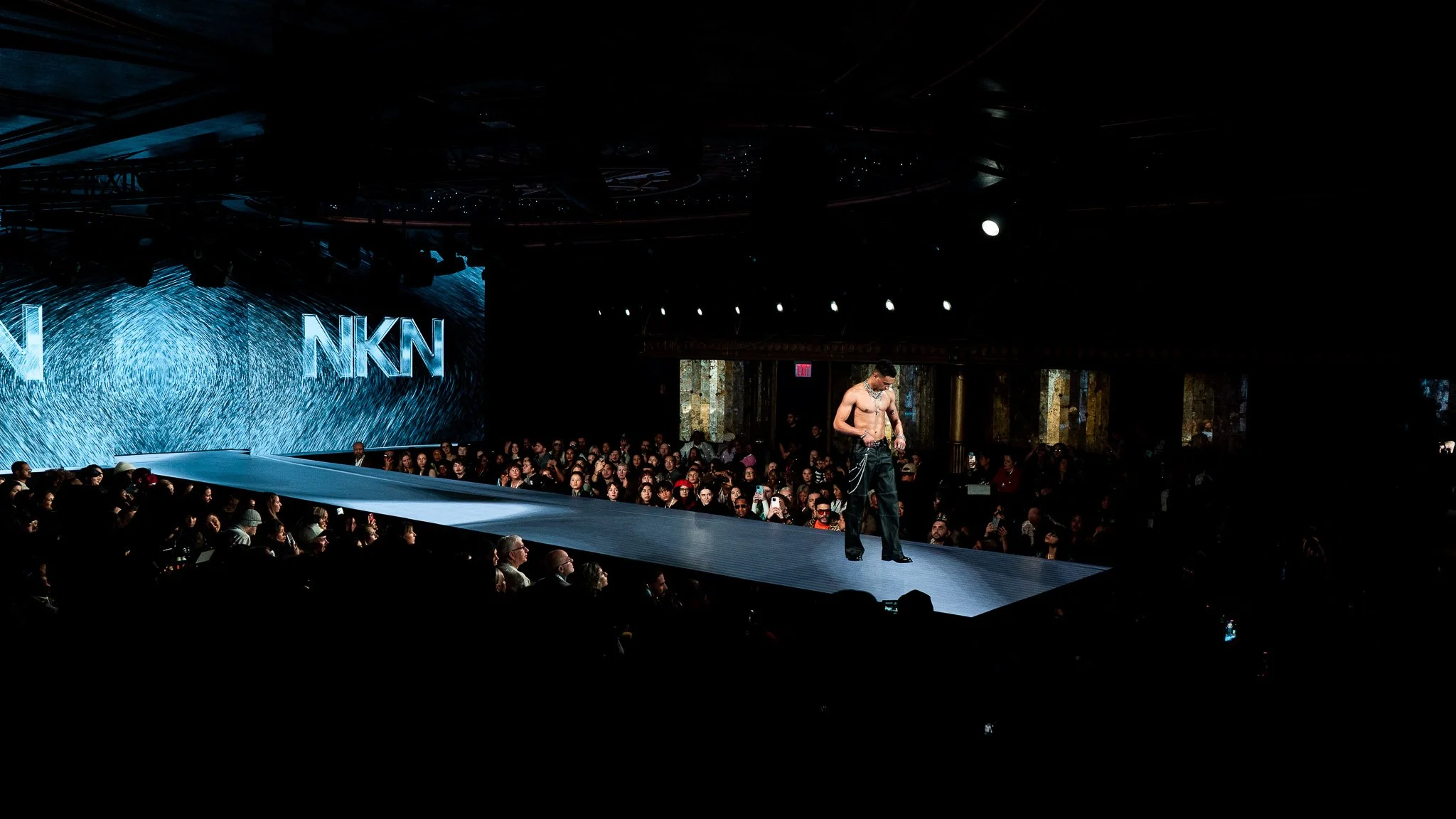 Fashion runway show with a male model walking on the catwalk, audience seated on both sides, large LED screen displaying abstract graphics and the letters 'NKN' in the background.