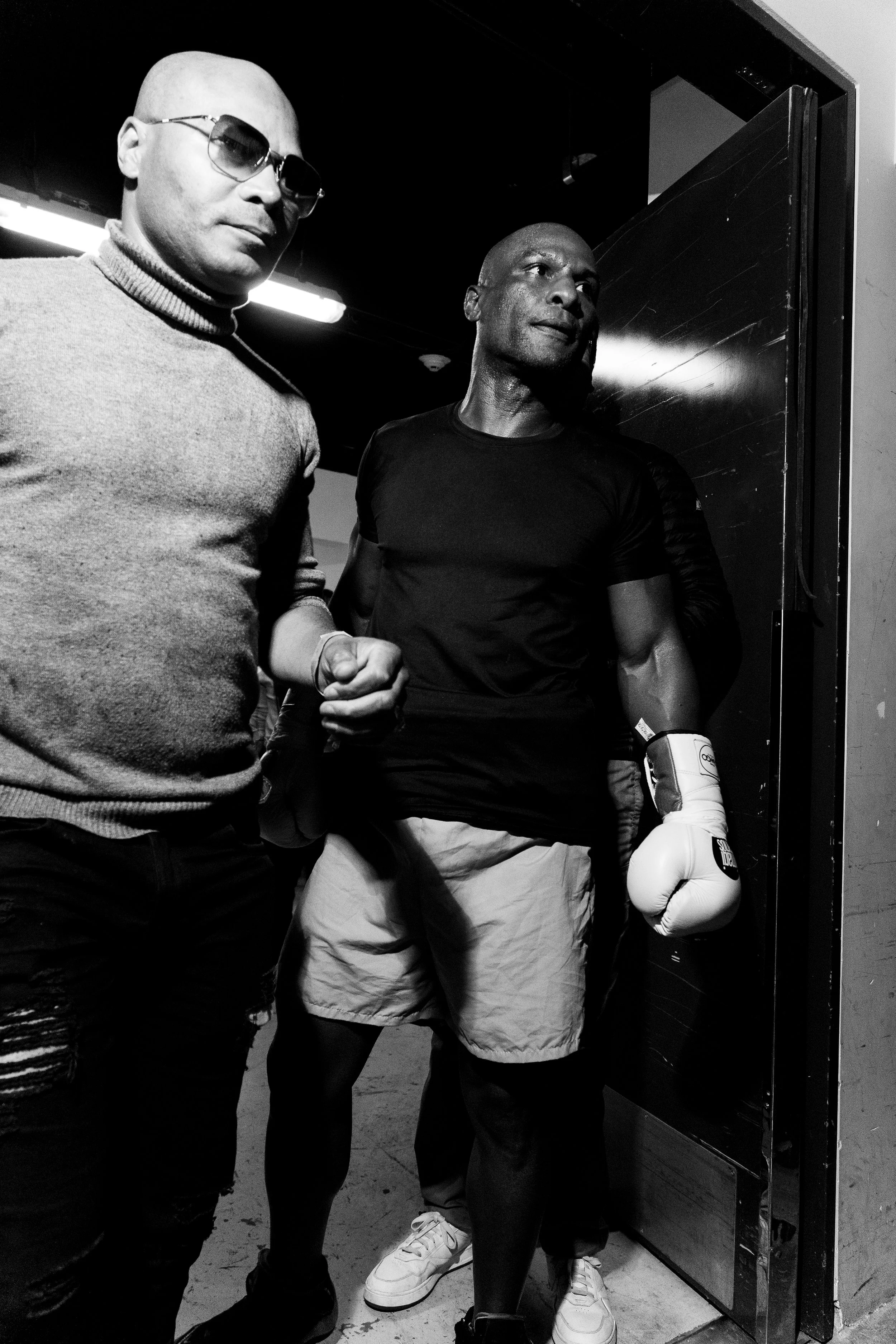 Two muscular men, one with glasses in a turtleneck sweater and the other in a black t-shirt and boxing gloves, are standing close to a metal door in a dimly lit room.