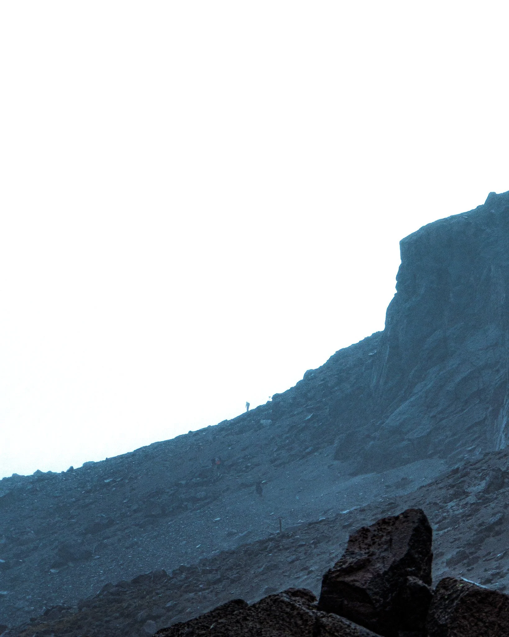 A mountain landscape with rocky terrain in the foreground and a large mountain peak in the background. The sky appears overexposed or white.