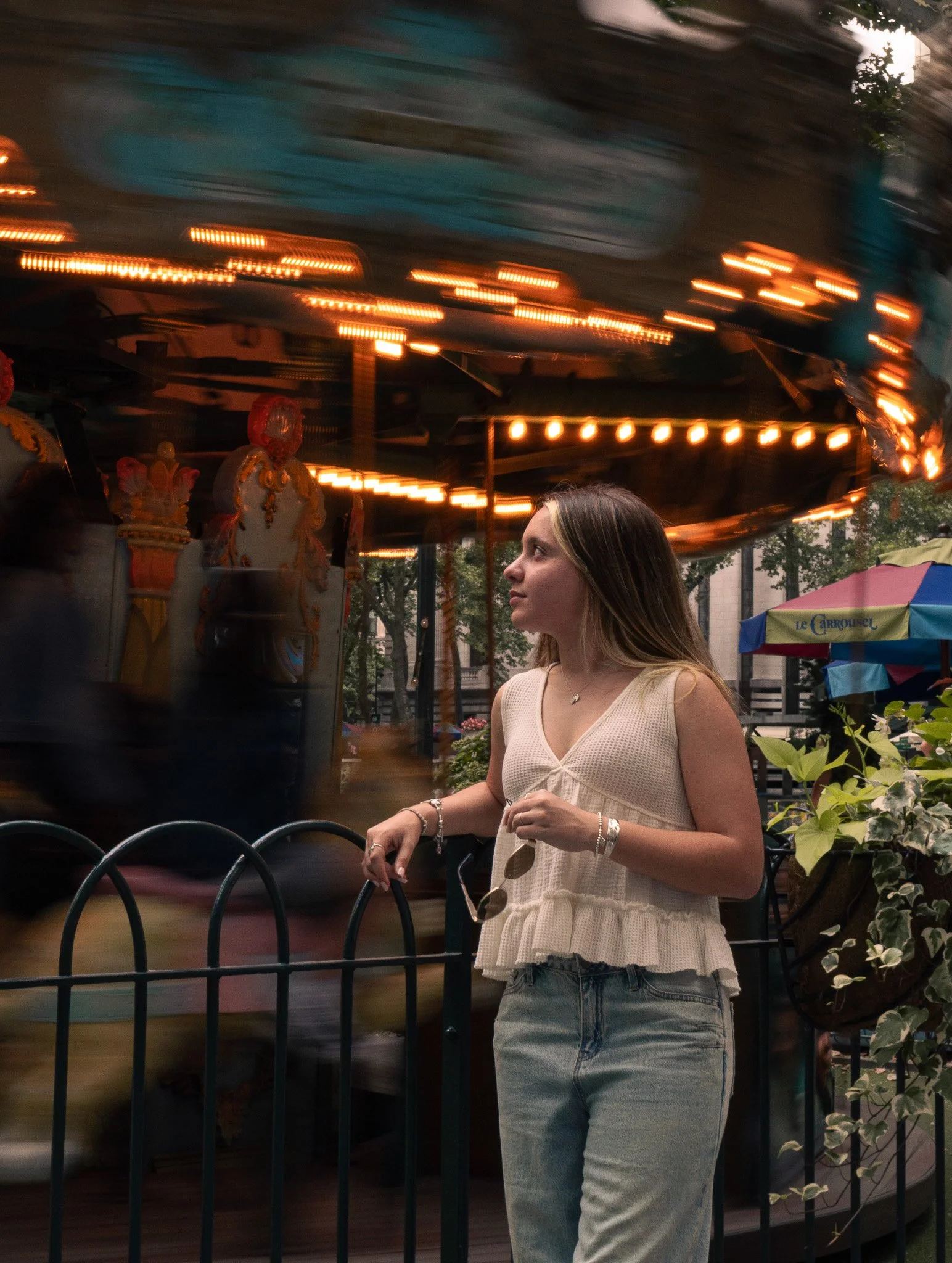 A young woman with long brown hair wearing a sleeveless beige top and blue jeans, standing outdoors near a black metal fence. Behind her, a carousel with illuminated lights is in motion, creating a blurred, swirling effect. Part of a colorful umbrell