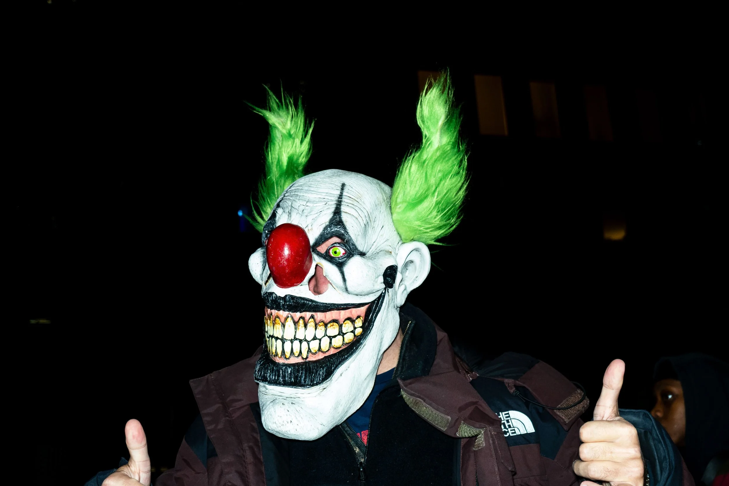 Person wearing a scary clown mask with green hair, red nose, and black and white face paint, giving a thumbs-up gesture.