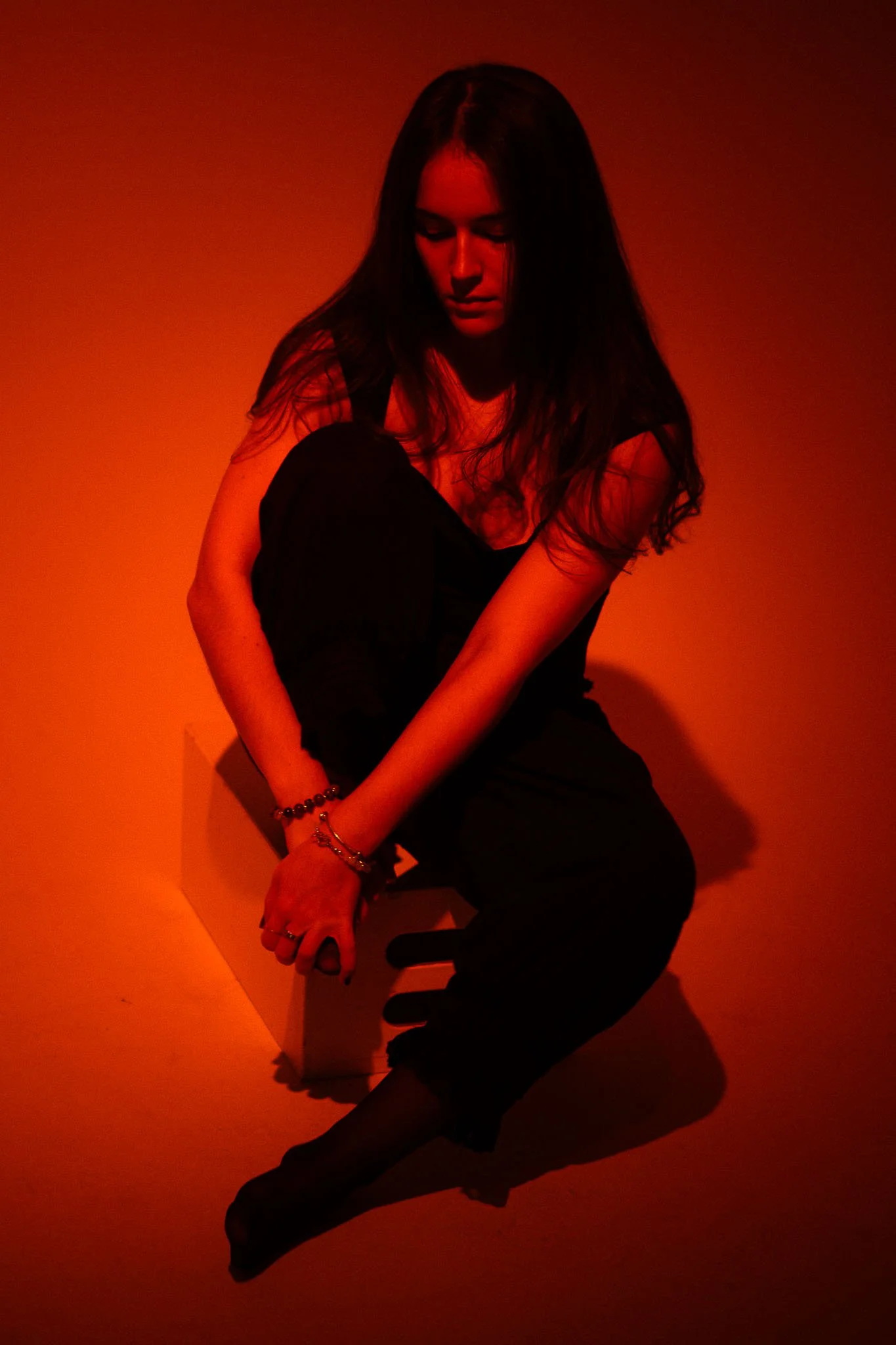 A woman with long dark hair is seated with her knees drawn up, wearing a black outfit and jewelry, illuminated by red lighting, against a dark background.