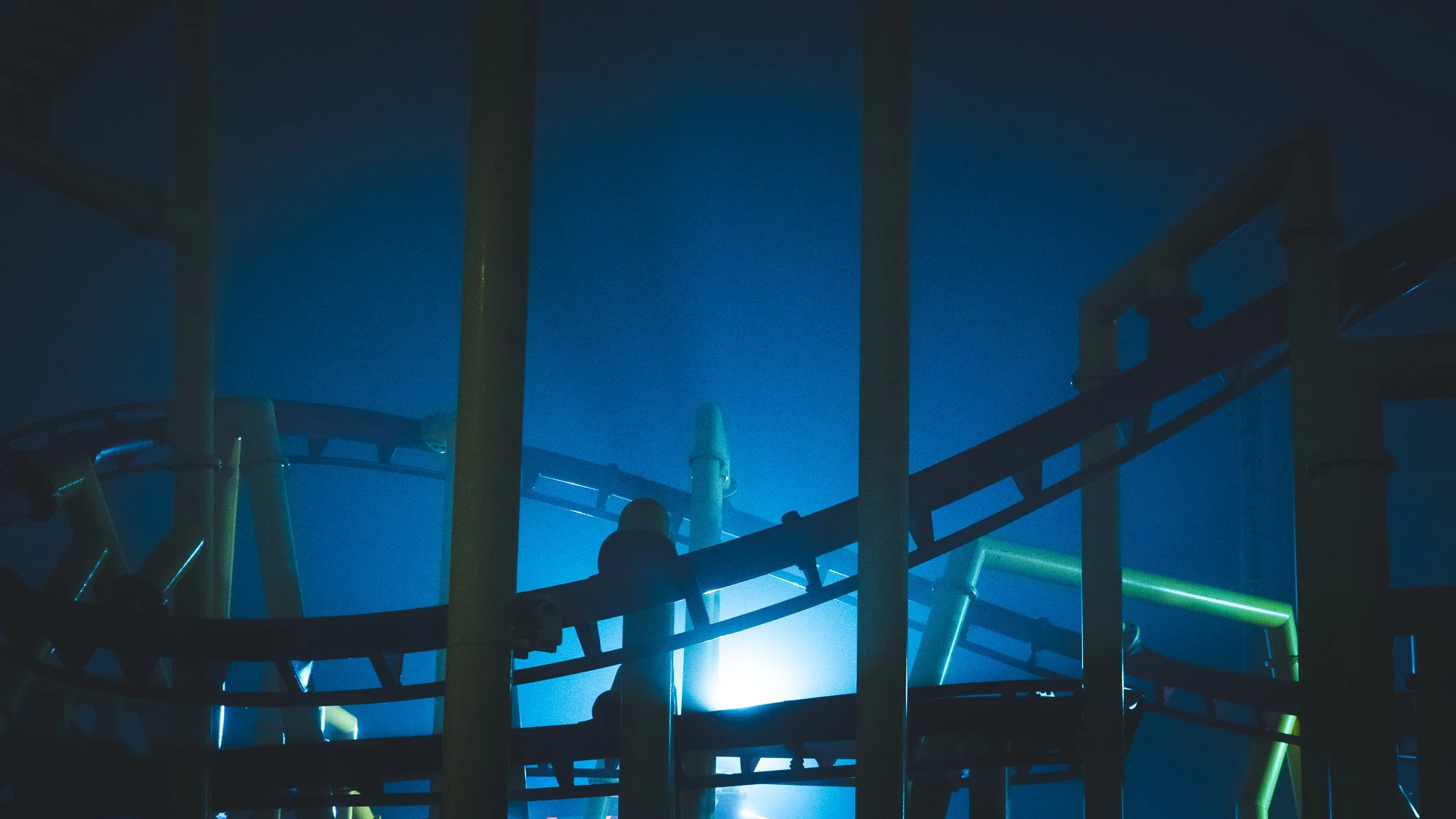 Dark amusement park ride with yellow and green pipes, illuminated by blue light, with a person sitting on the ride.