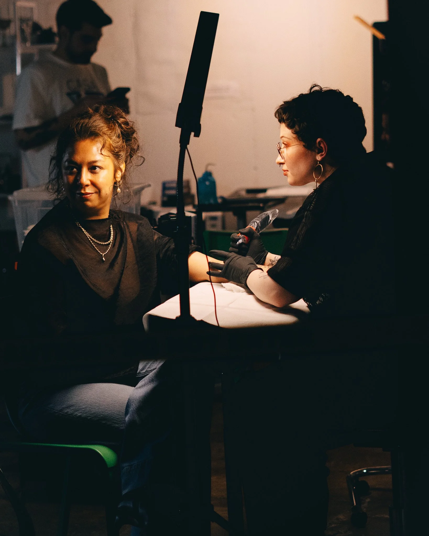 A person with short curly hair and glasses, sitting at a table with a tattooed arm, receiving a tattoo from a tattoo artist wearing gloves. The person in the background is using a phone.