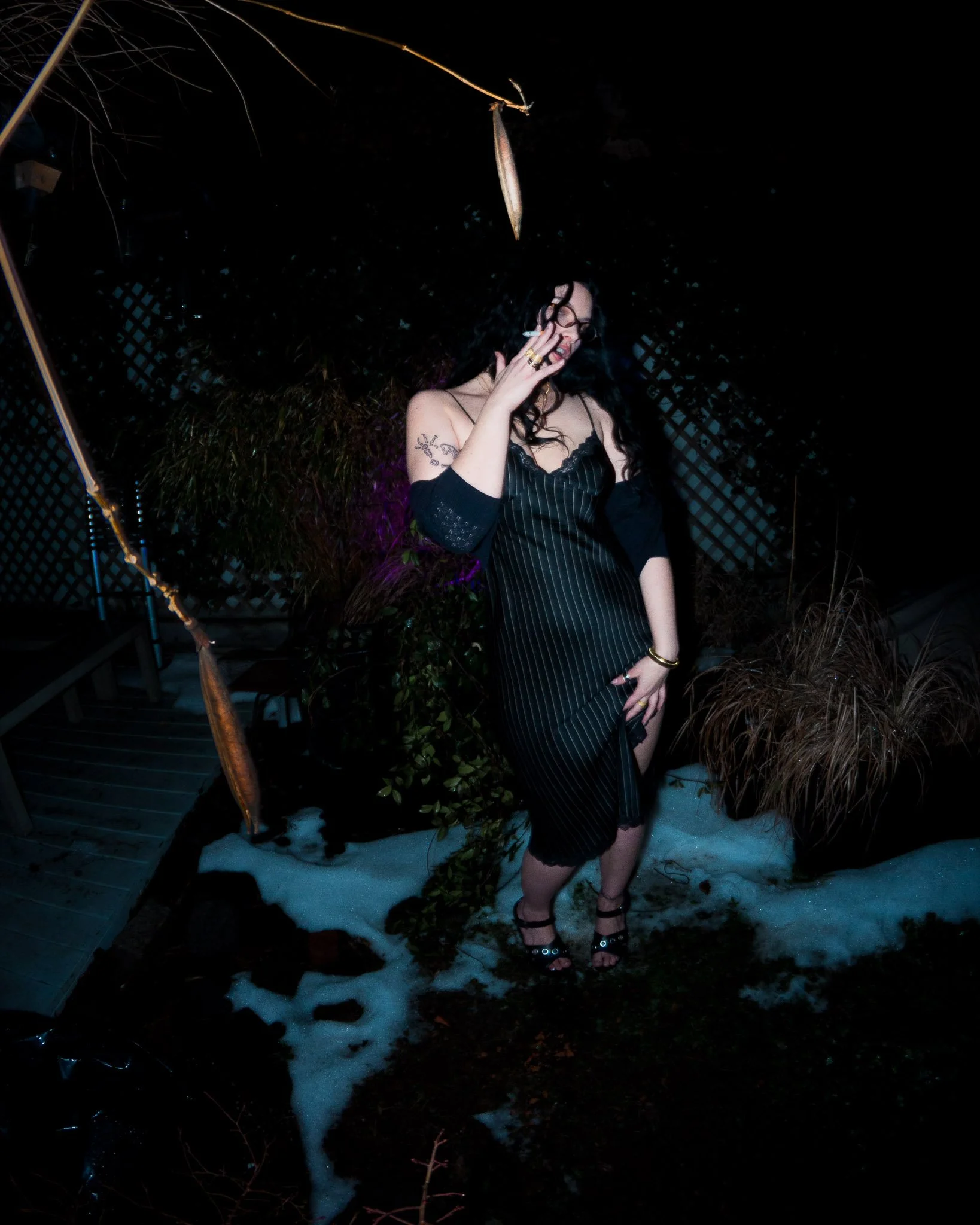 A woman with black, curly hair wearing glasses, a black off-shoulder dress with vertical stripes, and black high-heeled sandals, standing outdoors at night on snow-covered ground. She is smoking a cigarette and holding her hand to her face. The backg