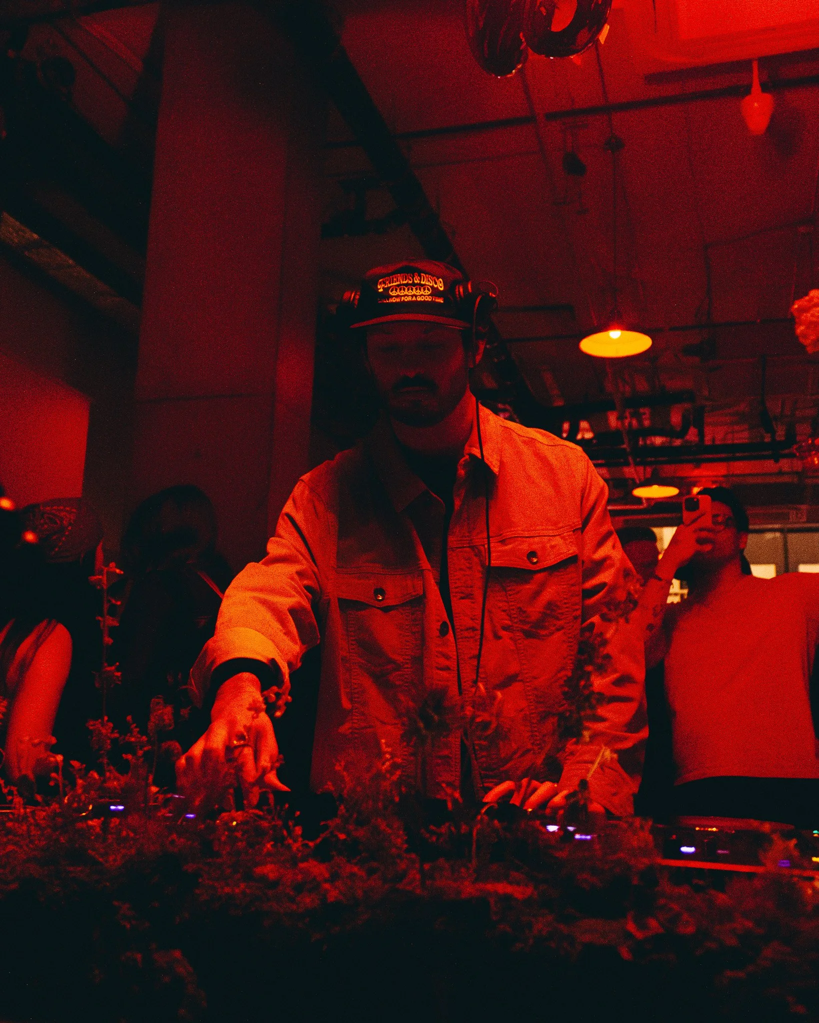 A person wearing a cap with the words "Friends & Disco" and an emoji pattern is DJing at a venue with red lighting, standing behind a DJ setup with record turntables.