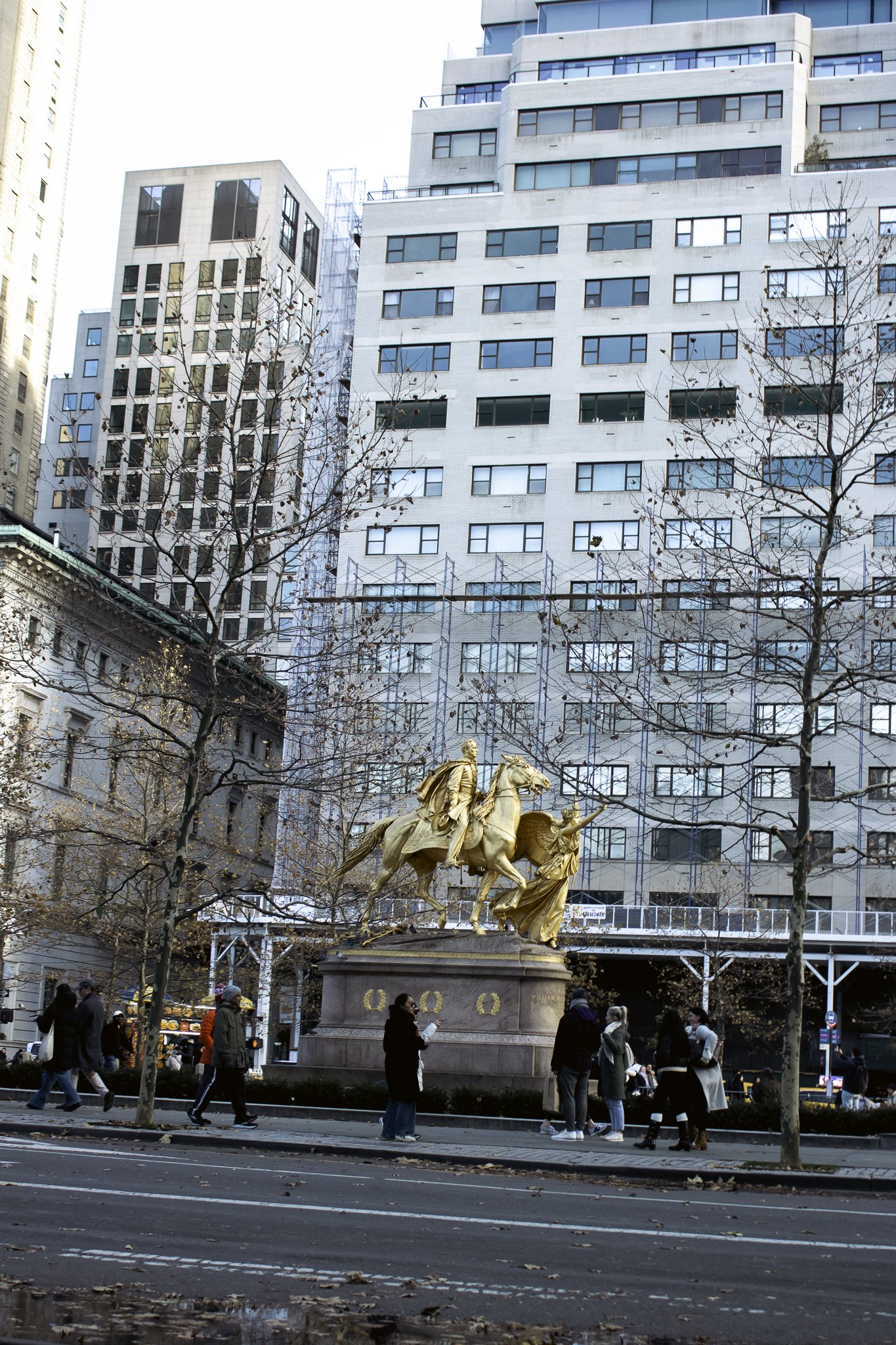 Gold sculpture of a person on horseback and another person walking beside them, located in an urban city square with tall buildings and leafless trees surrounding it. Pedestrians walk along the street in the foreground.
