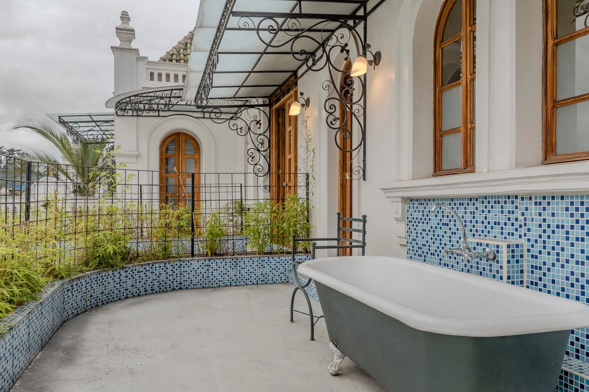 A rooftop patio with a clawfoot bathtub next to a blue mosaic tiled wall, surrounded by a black metal fence and wooden window frames, with a cloudy sky in the background.