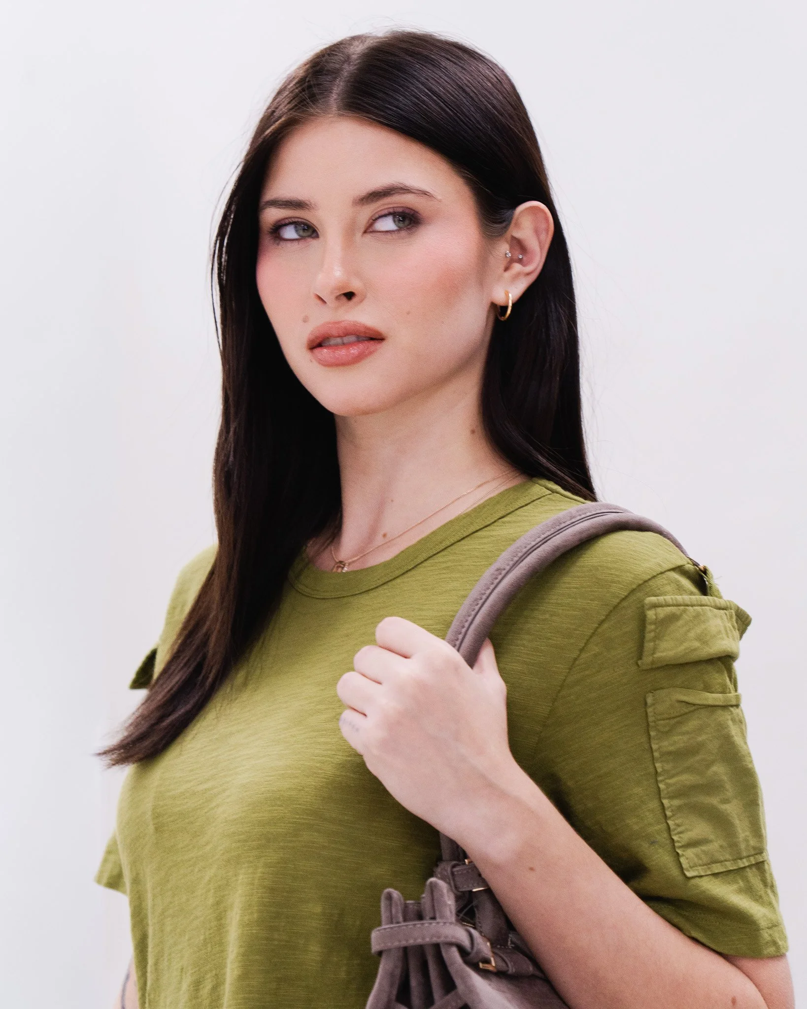 A woman with long dark hair, wearing a green shirt, and with multiple earrings in her right ear, holding a bag strap with her left hand, looking to the side against a plain, light background.