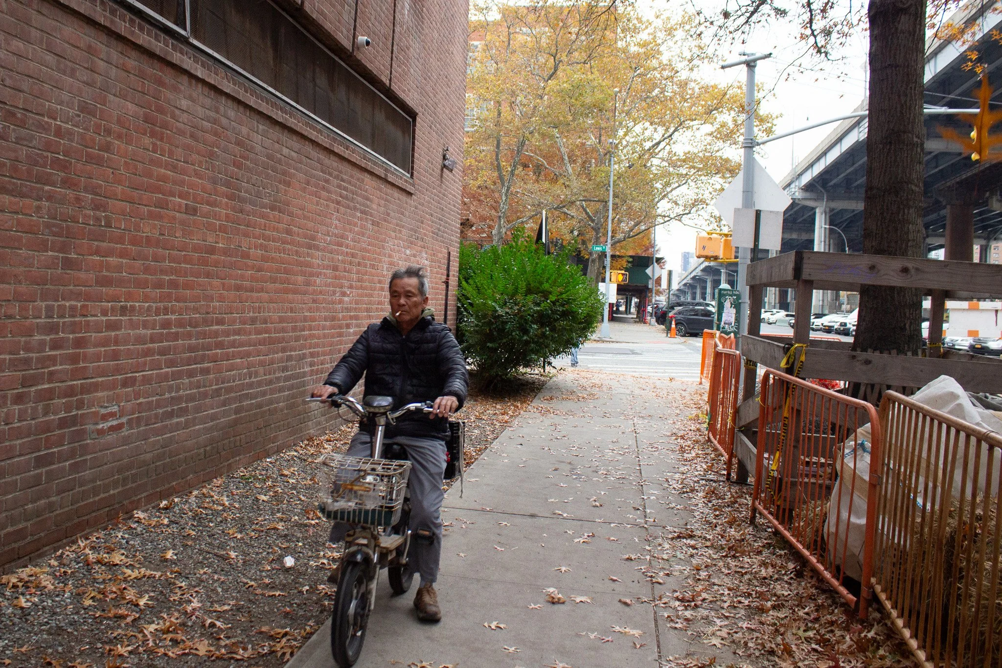 A man in a black jacket riding a bicycle on a city sidewalk during autumn, with fallen leaves on the ground, a brick wall on the left, and trees and street scene in the background.