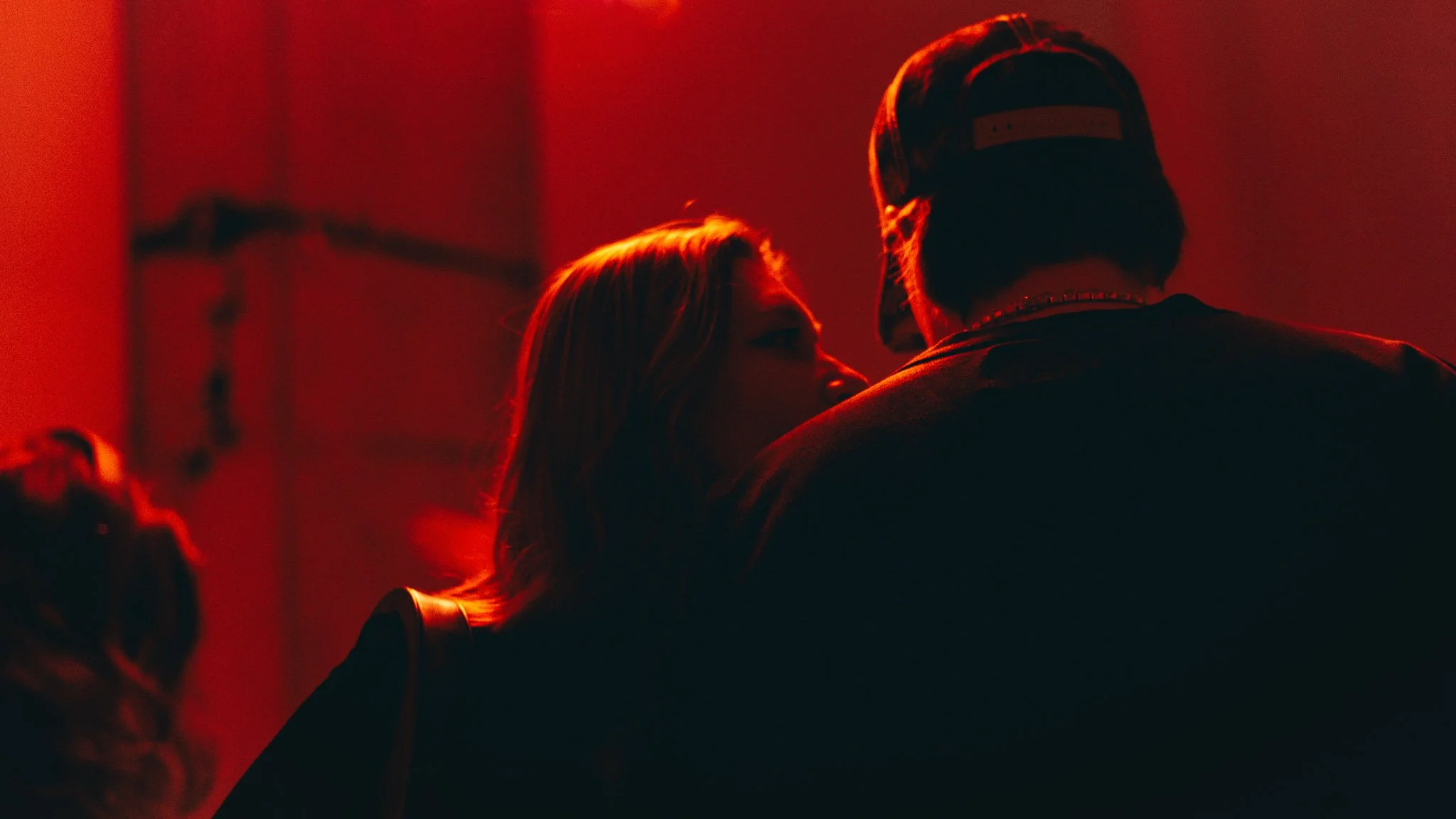 A woman and a man close together in a dimly lit environment with red lighting, possibly a club or party setting.