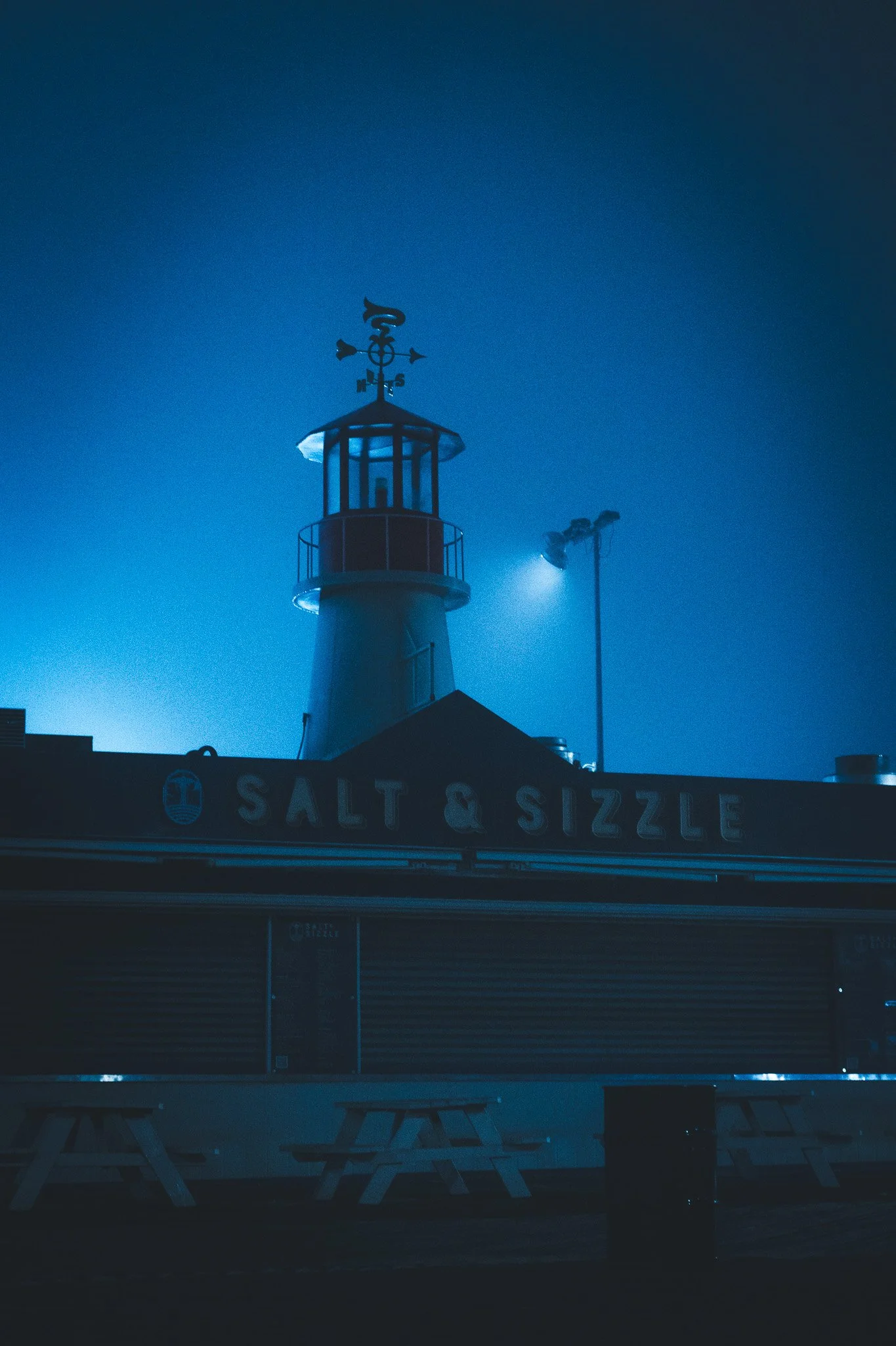 Nighttime scene of a lighthouse with a weather vane on top, illuminating with blue light, and a restaurant called 'Salt & Sizzle' with closed shutters and outdoor picnic tables in front.