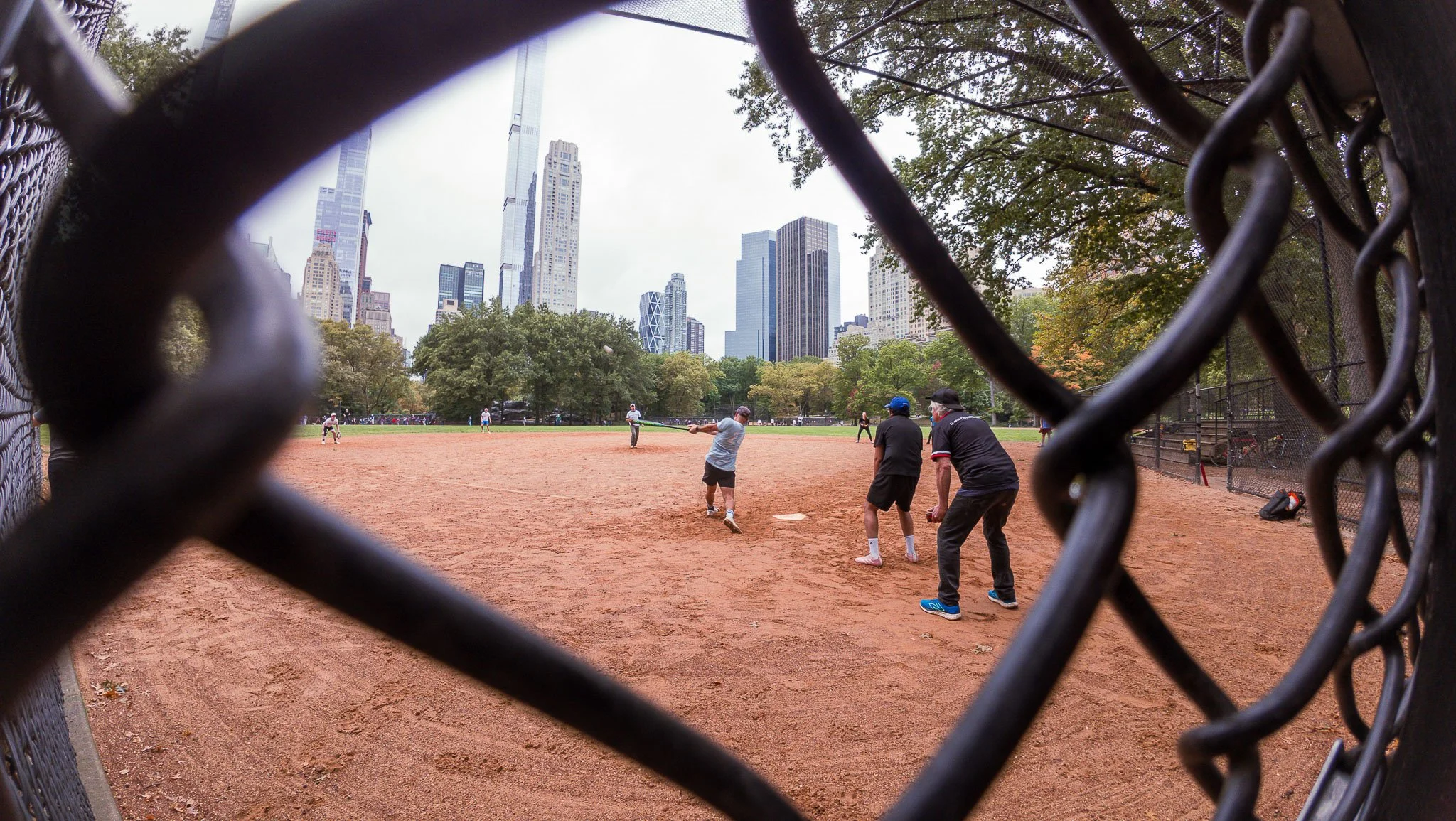 People playing baseball in a park with a city skyline in the background, viewed through a chain-link fence.