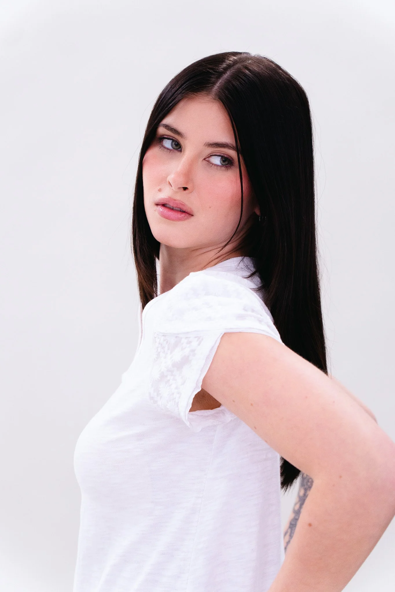 A woman with long black hair wearing a white shirt, looking over her shoulder at the camera, standing against a plain white background.
