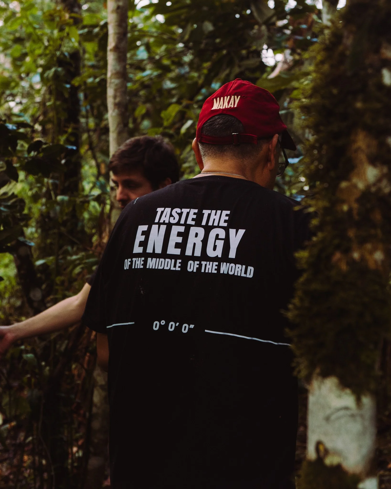 Two people in a forest, one wearing a red cap with 'MAKAY' written on it and a black shirt with white text about energy, the other partially visible.
