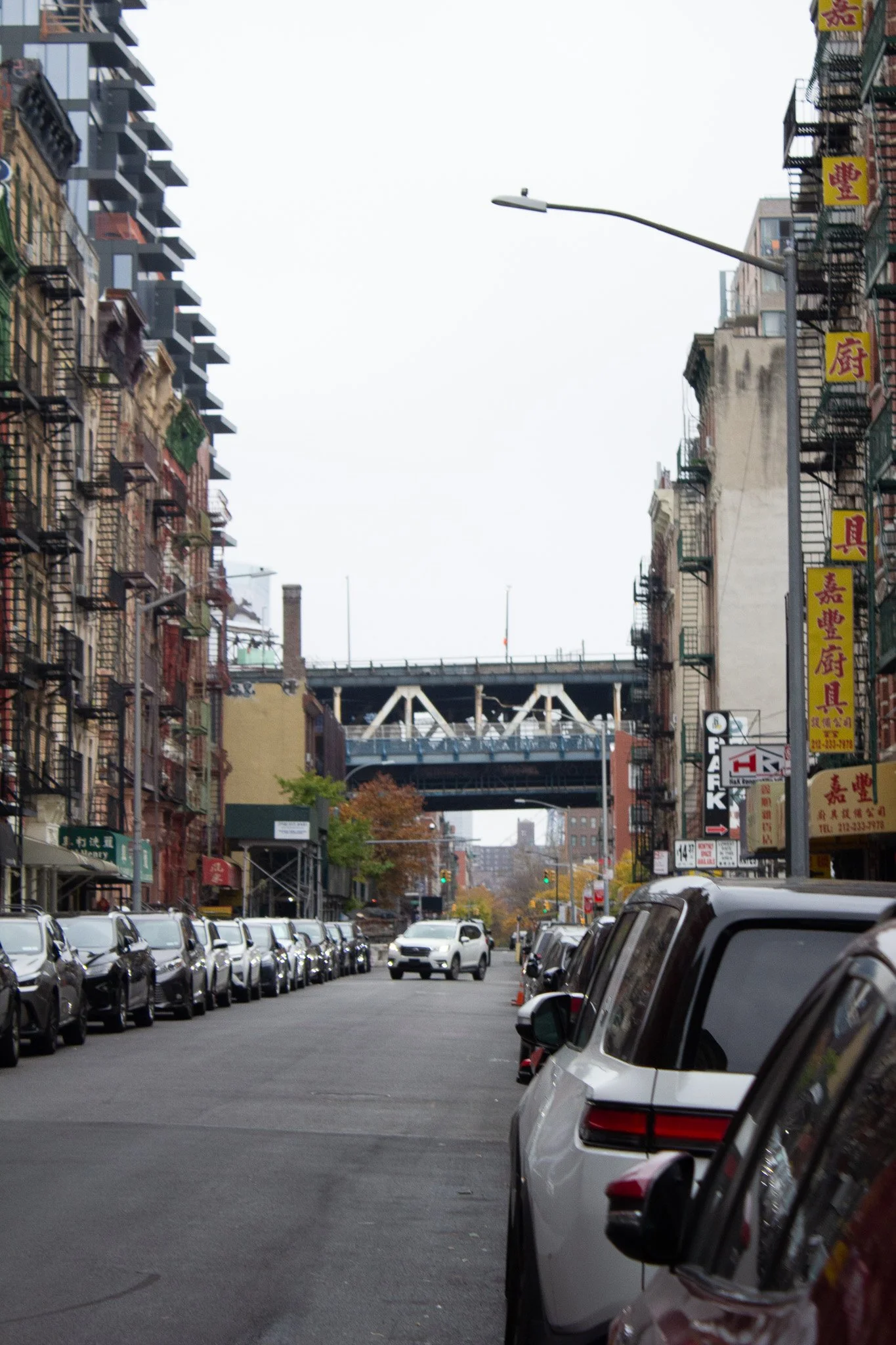 A city street scene with parked cars lining both sides, buildings with fire escape ladders and Chinese signage on the right, and a street intersection with traffic lights. A bridge is visible overhead in the background.