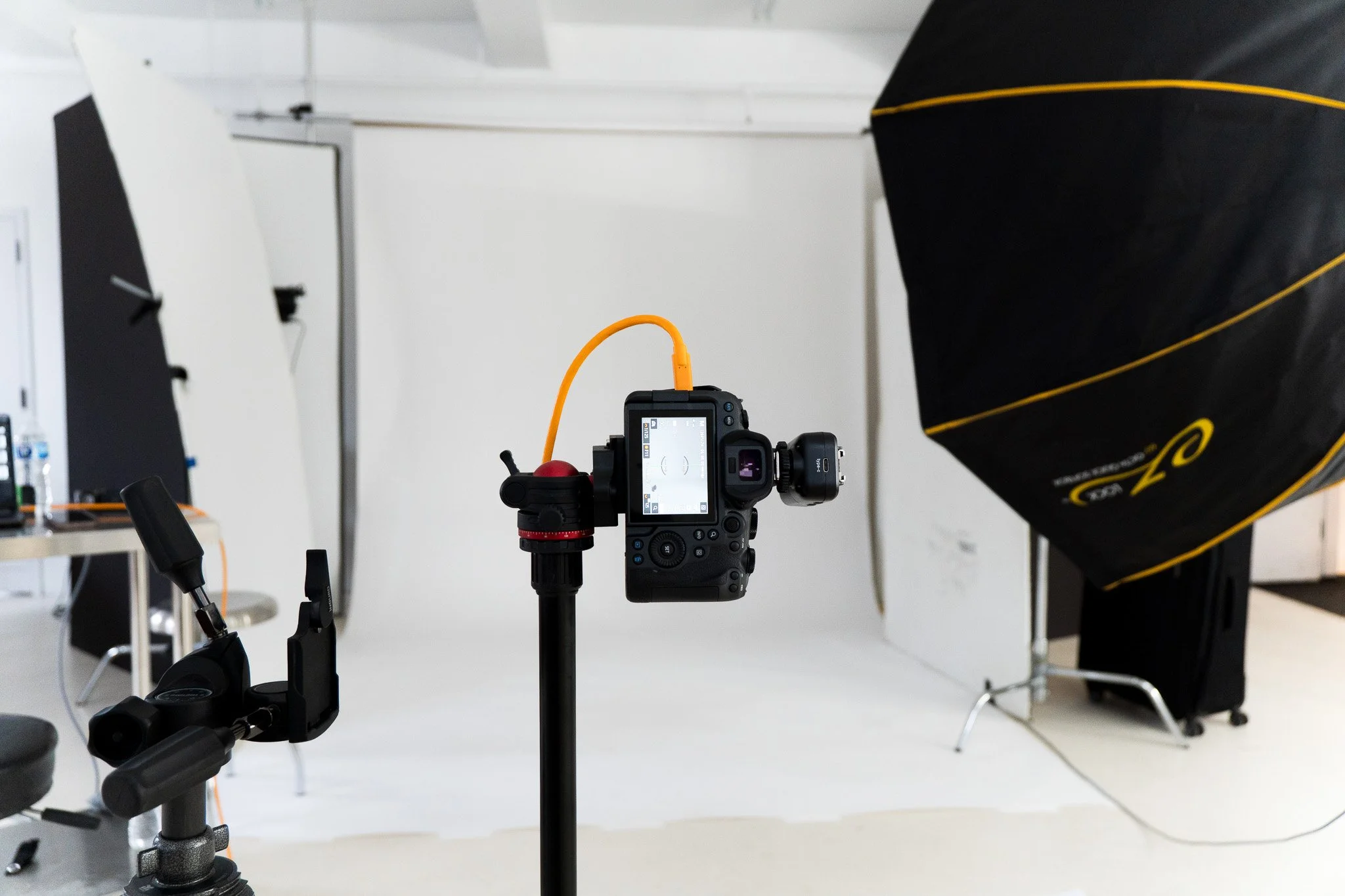 Studio setup with a DSLR camera on a tripod, connected to a computer or controller, aimed at a backdrop with a white wall and black fabric diffuser.