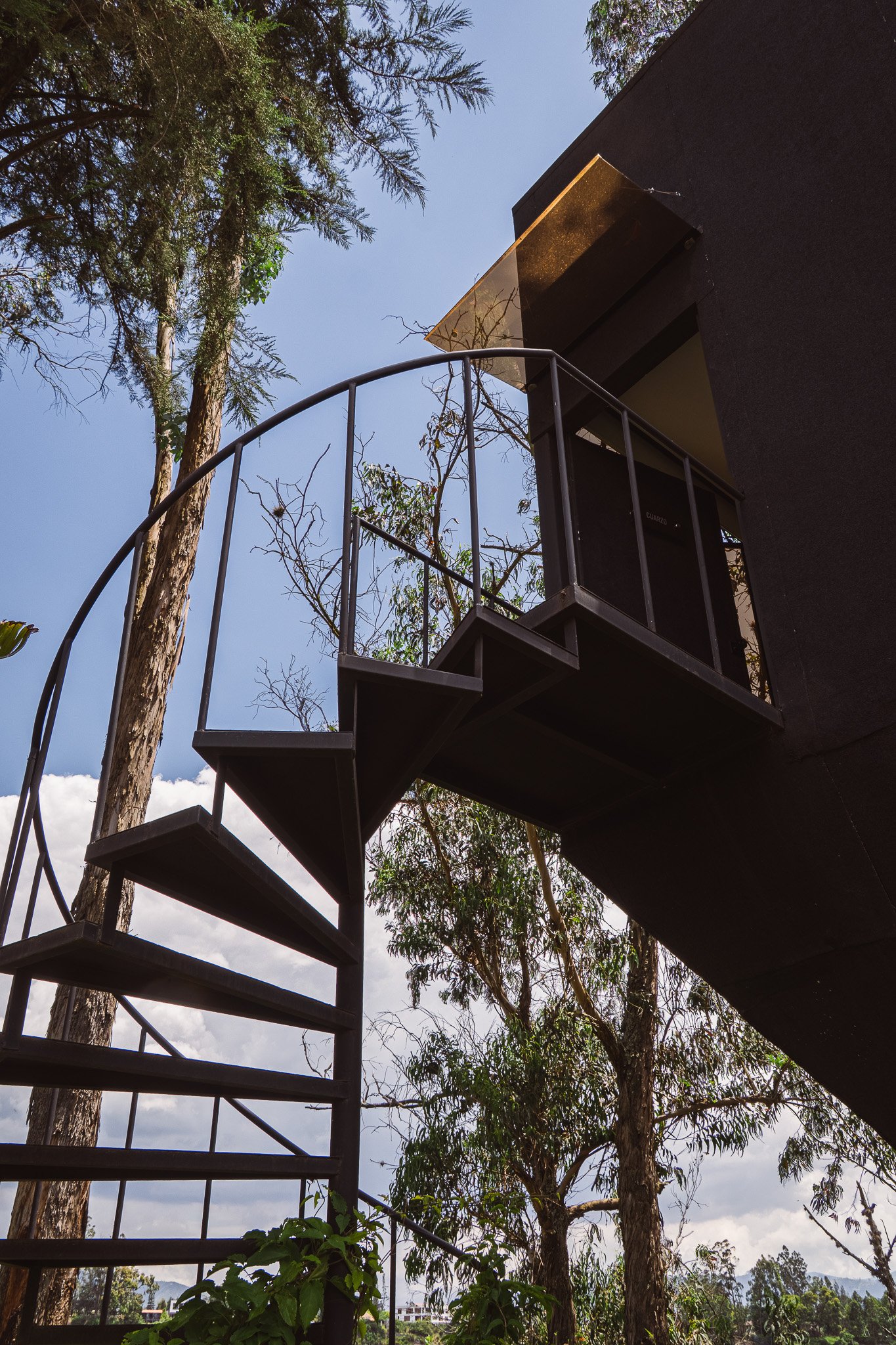 Black metal staircase with a small balcony and railing, set against a building with dark walls, surrounded by tall trees with green foliage, under a partly cloudy sky.