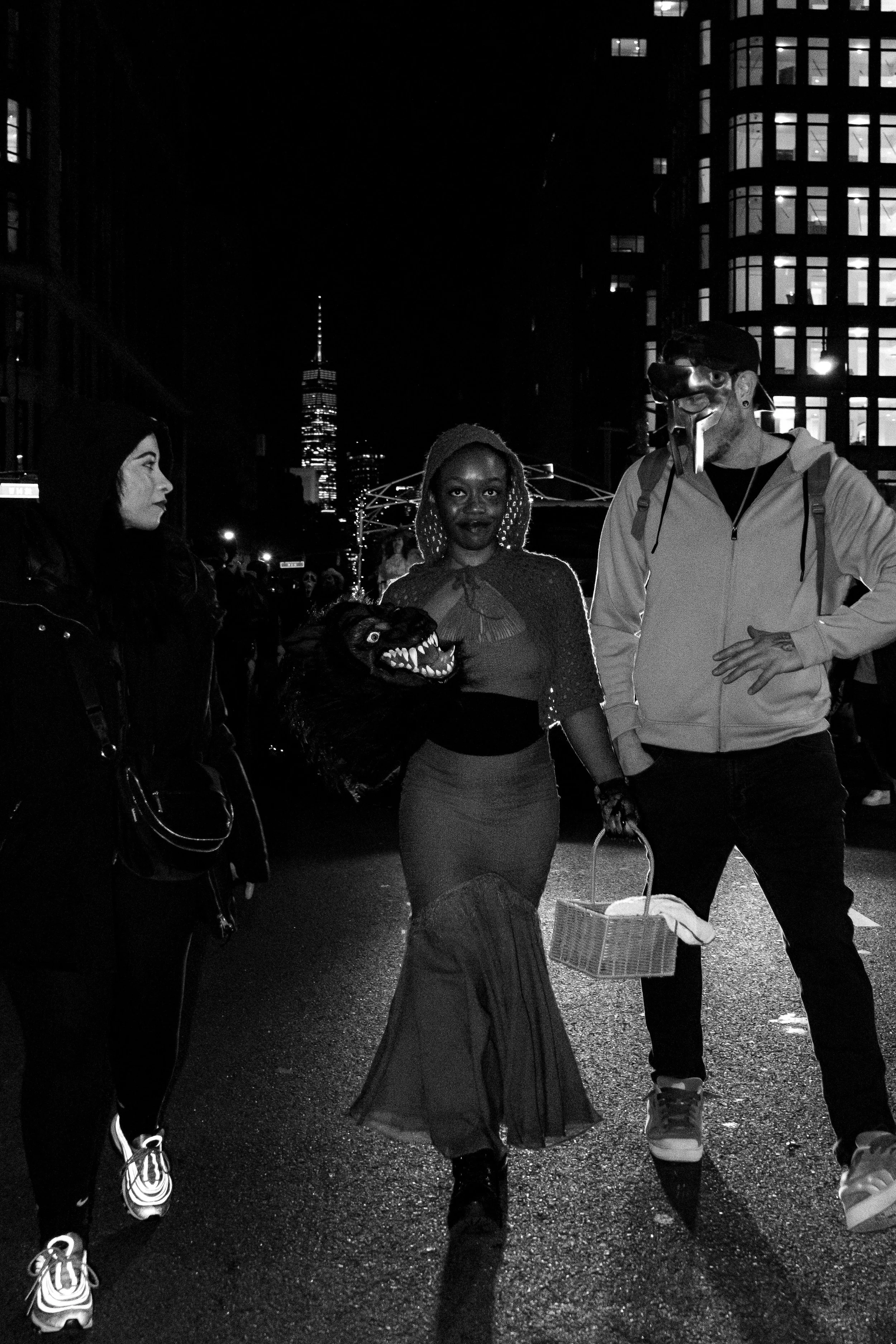 People dressed in costumes walking on a city street at night, with tall buildings in the background, including the One World Trade Center in New York City.