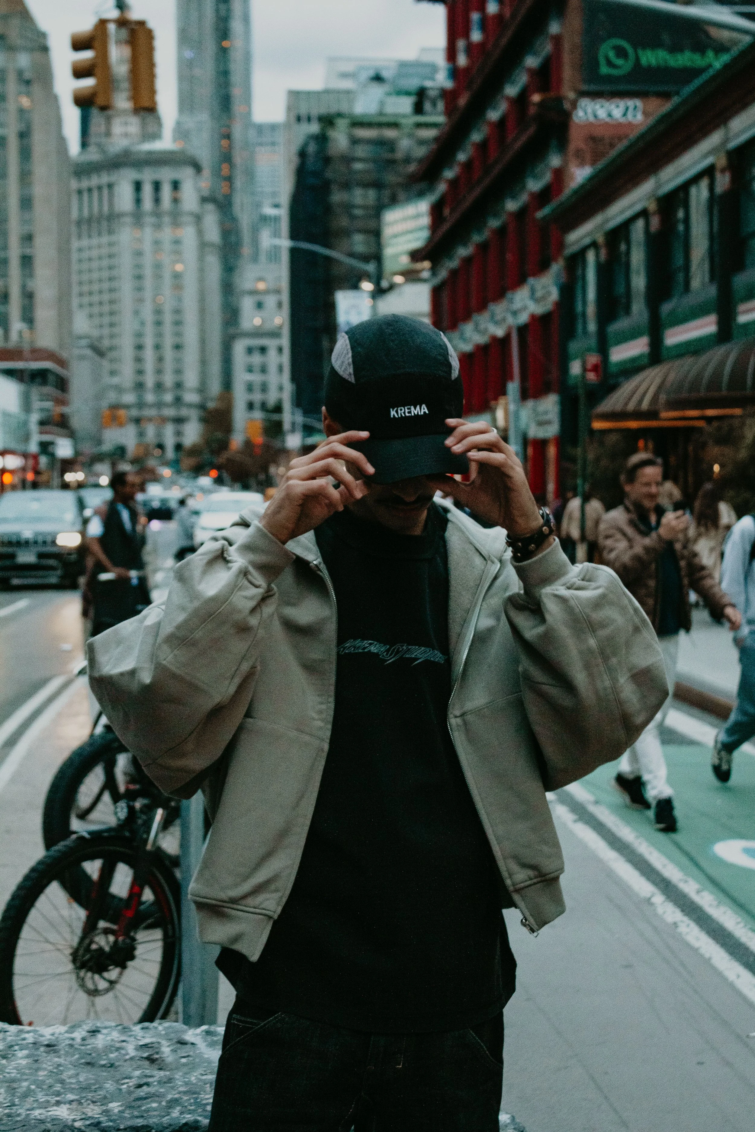 Man in black cap with 'Krema' logo adjusting it on busy city street with tall buildings and people walking around.