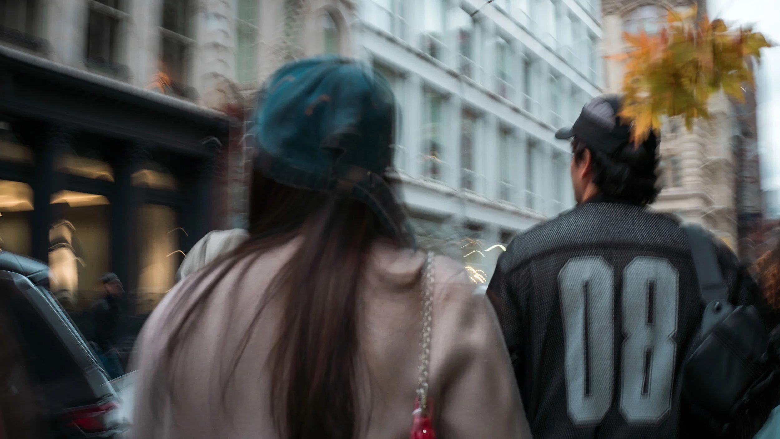 A woman and a man walking on the street, seen from behind, with blurred buildings and trees in the background.