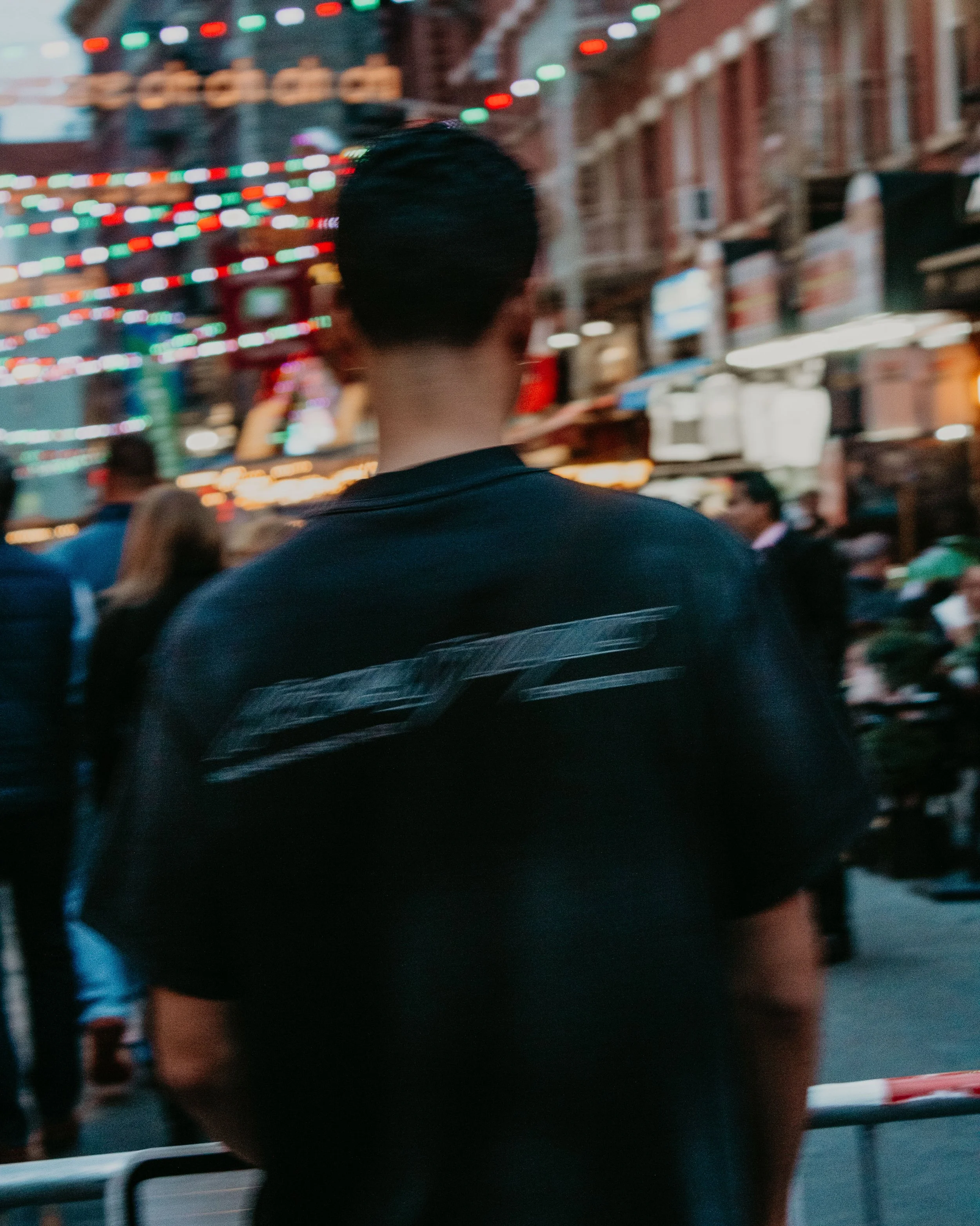 Back of a person with short black hair wearing a black jacket, standing in a busy street with colorful string lights, people, and storefronts in the background.