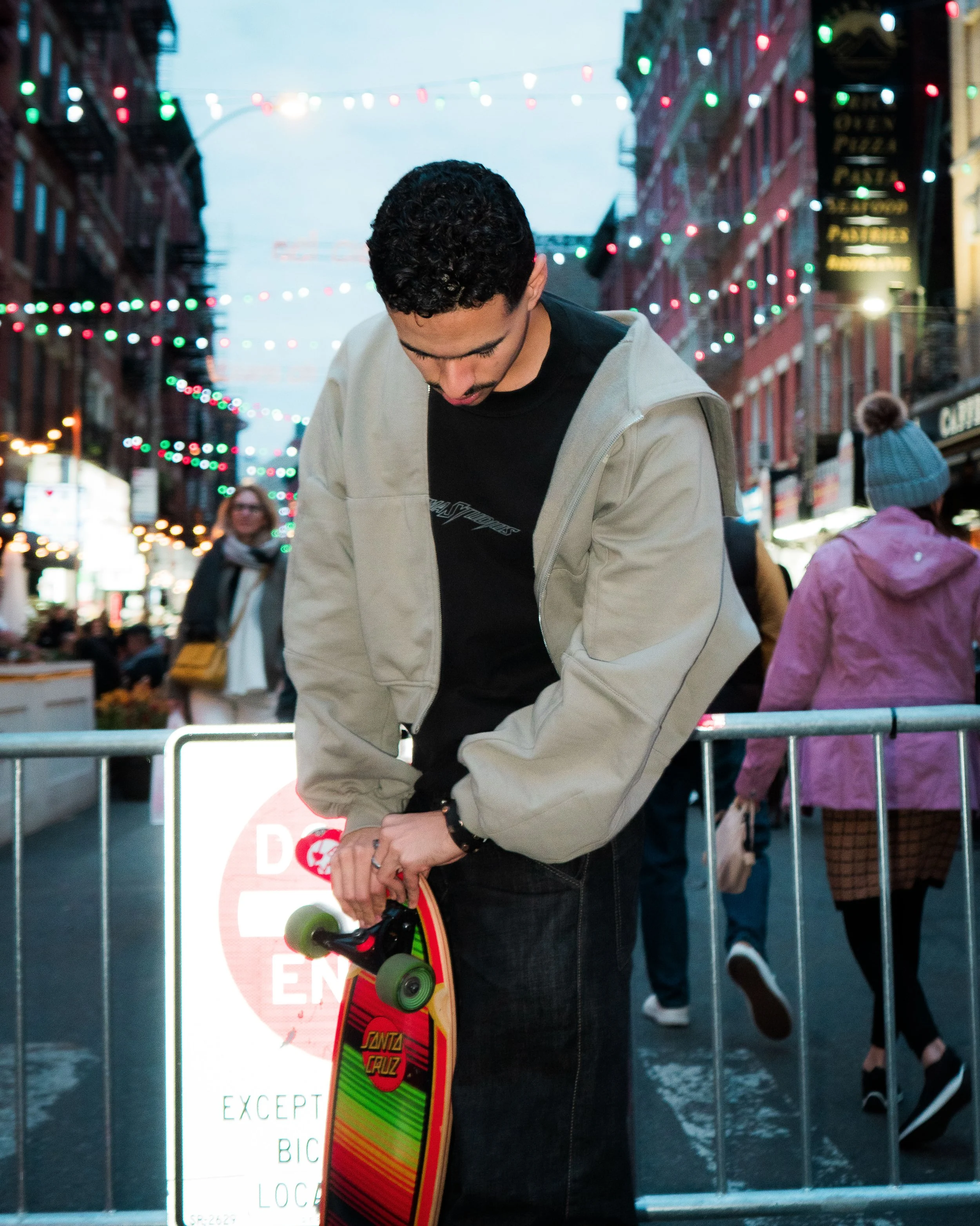 A young man with dark curly hair adjusting his skateboard at a busy city street with festive overhead string lights. He wears a gray jacket over a black shirt, standing near a barrier, with other pedestrians and buildings in the background.