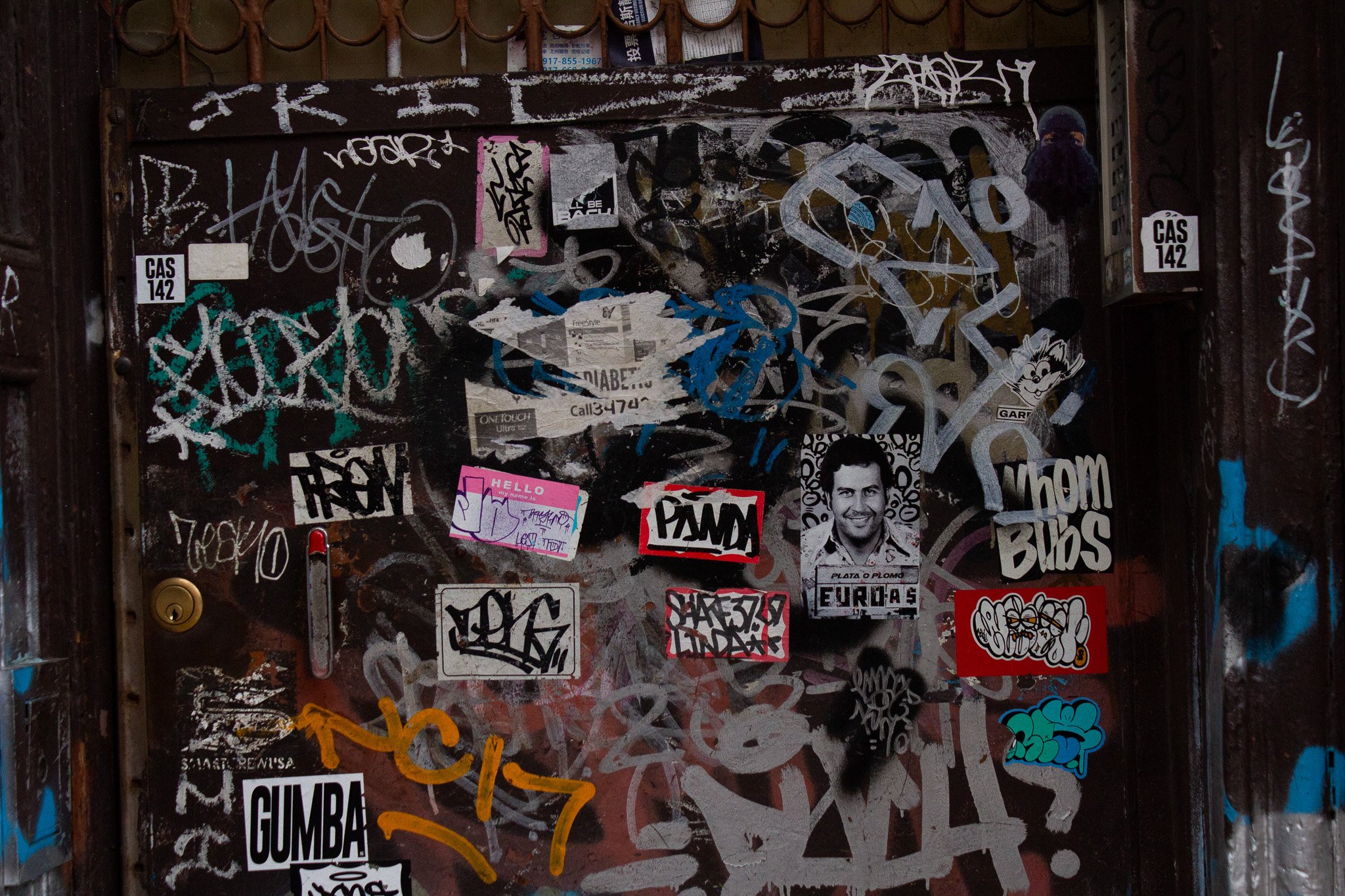 A wall covered with various graffiti, stickers, and posters. Includes tags, scribbles, and a black and white photo of a smiling man labeled 'EURAS'.