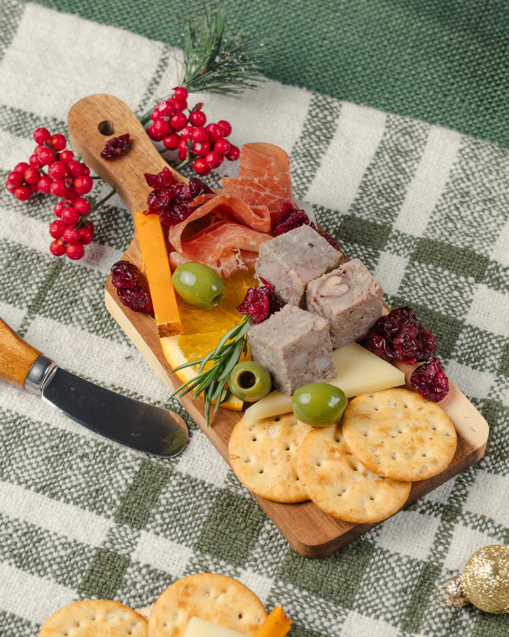 Close-up of a cheese and charcuterie board with crackers, green olives, sliced cured meat, cheese, dried cranberries, and fresh rosemary on a wooden tray, on a green and white checkered tablecloth.