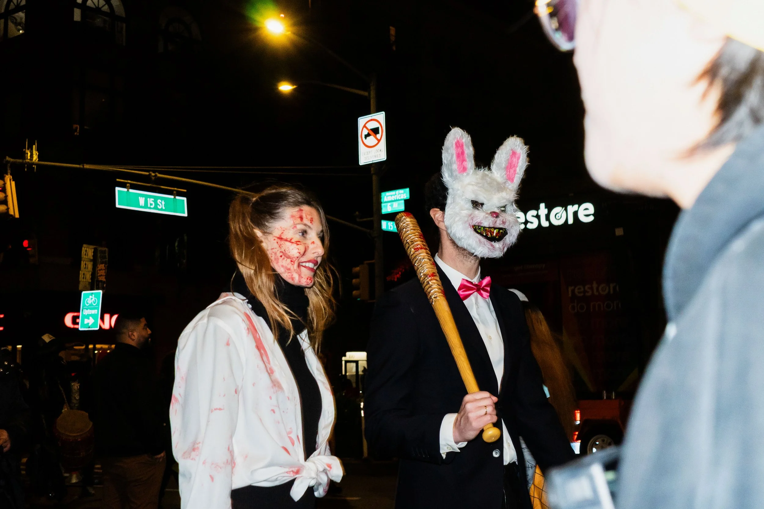 People dressed in Halloween costumes on a city street at night, including a woman with fake blood on her face and a man in a white rabbit mask and tuxedo holding a bat, engaging in conversation.