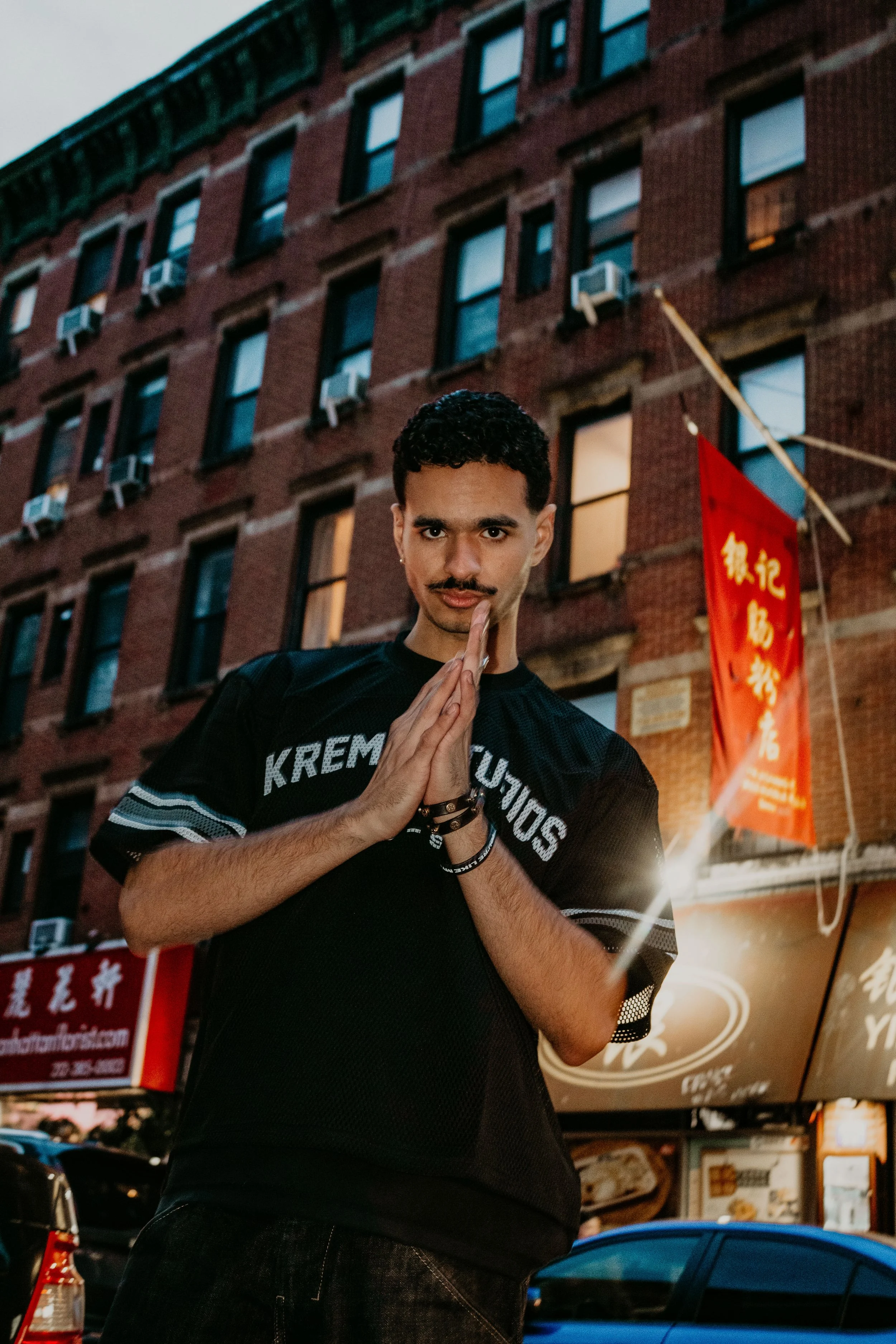 A young man with dark curly hair, a mustache, and a goatee posing with palms pressed together in a prayer-like gesture, standing in front of a brick building with multiple windows and air conditioning units, during dusk or evening.