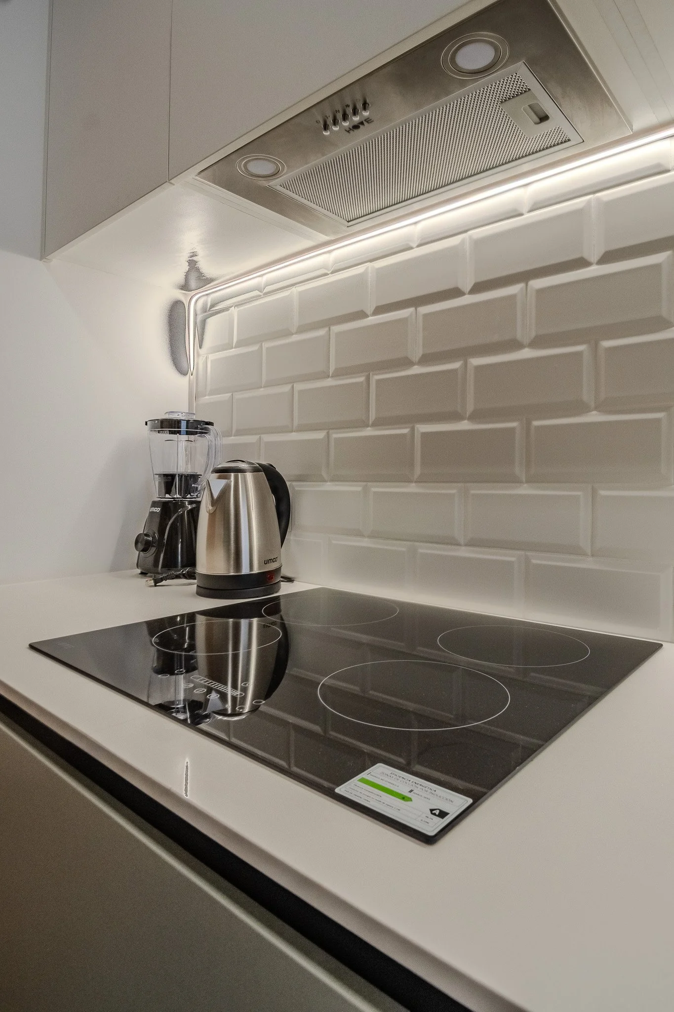 Modern kitchen with white cabinetry, a black electric cooktop, a stainless steel kettle, a blender, white subway tile backsplash, and under-cabinet lighting.