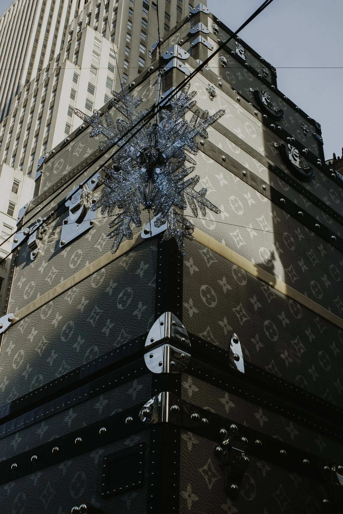 View of a corner of a Louis Vuitton patterned luggage piece with a decorative snowflake ornament hanging on its corner, set against tall buildings and a clear sky.