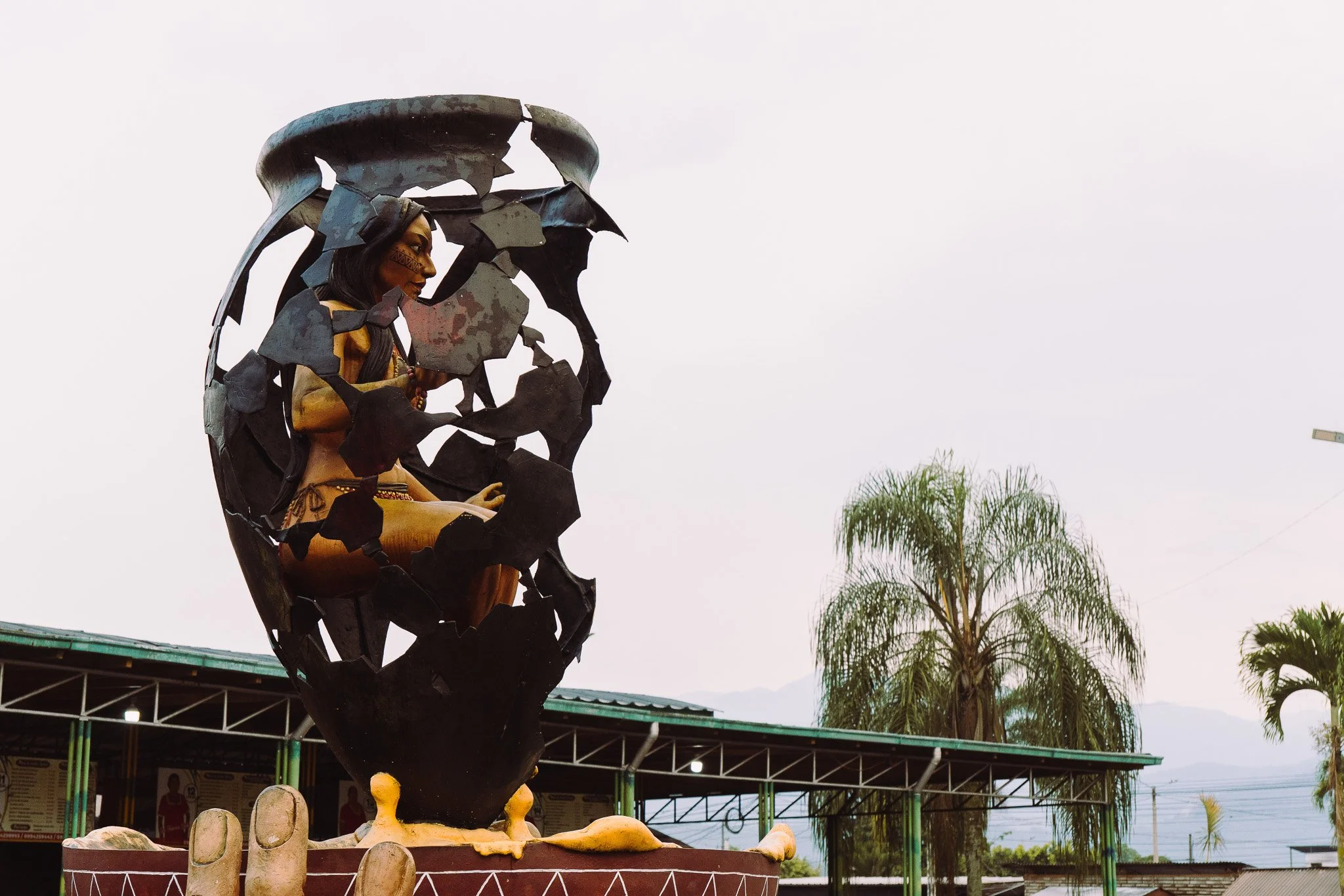 A sculpture of a woman with traditional face paint and jewelry, sitting inside an open metal structure shaped like a cup or bowl. The sculpture is set outdoors with palm trees and a cloudy sky in the background.