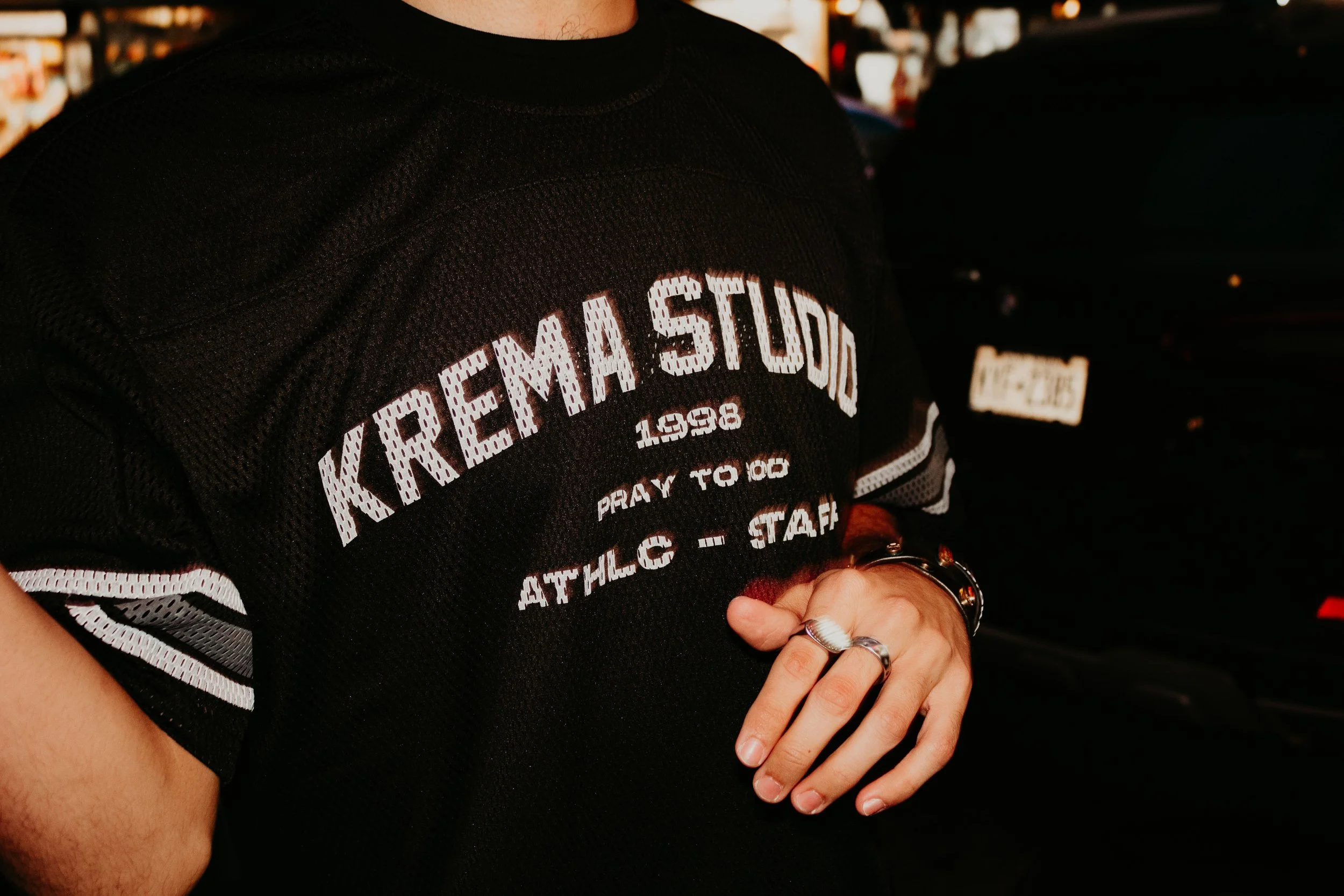 Person wearing a black sports jersey with white lettering that says 'KREMA STUDIOS 1998 PRAY TO GOD ATHLEC - STAFF' and rings on their fingers, standing outdoors near cars.