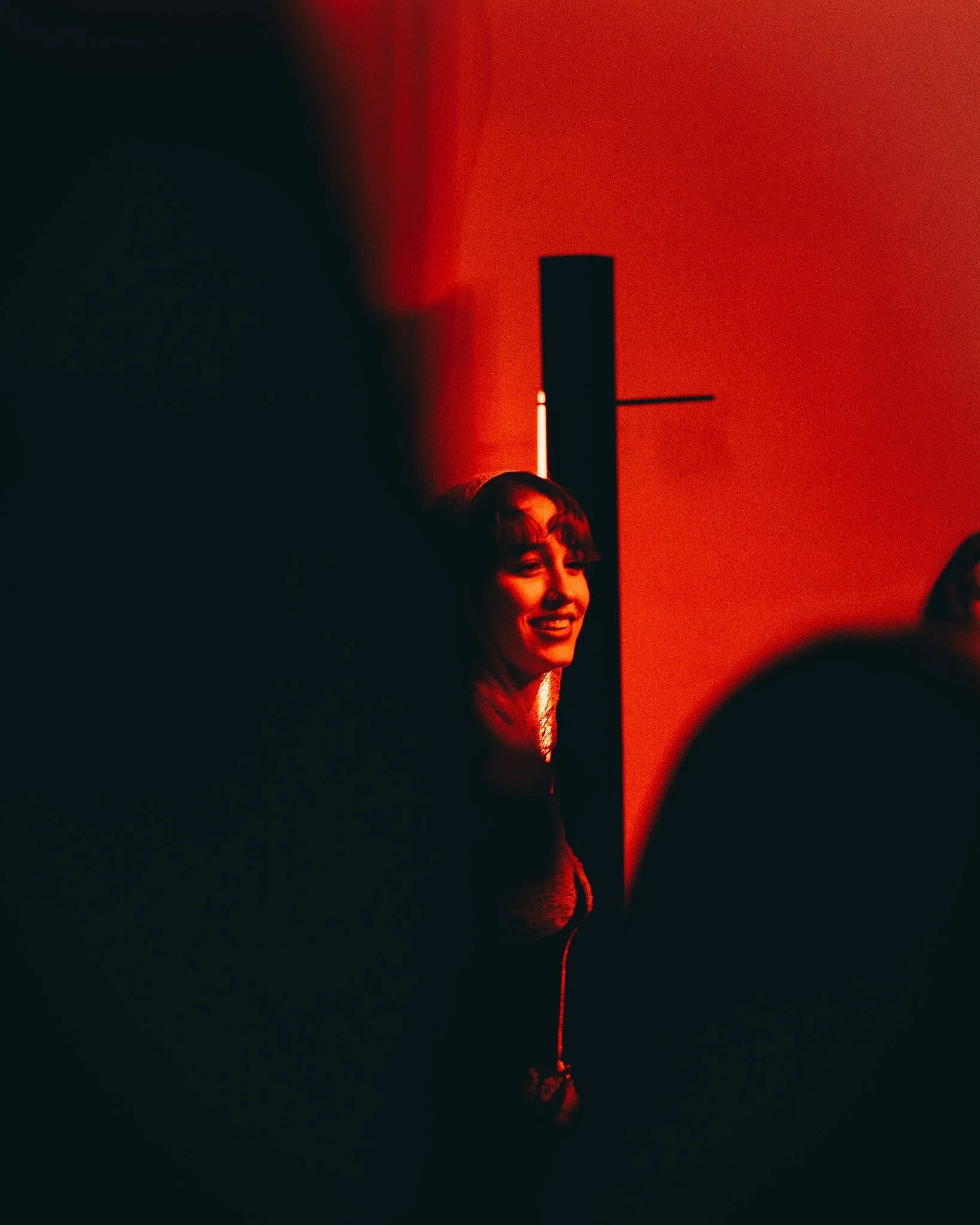 Smiling woman with long hair, lit by red lighting, standing near a black object on a red wall.