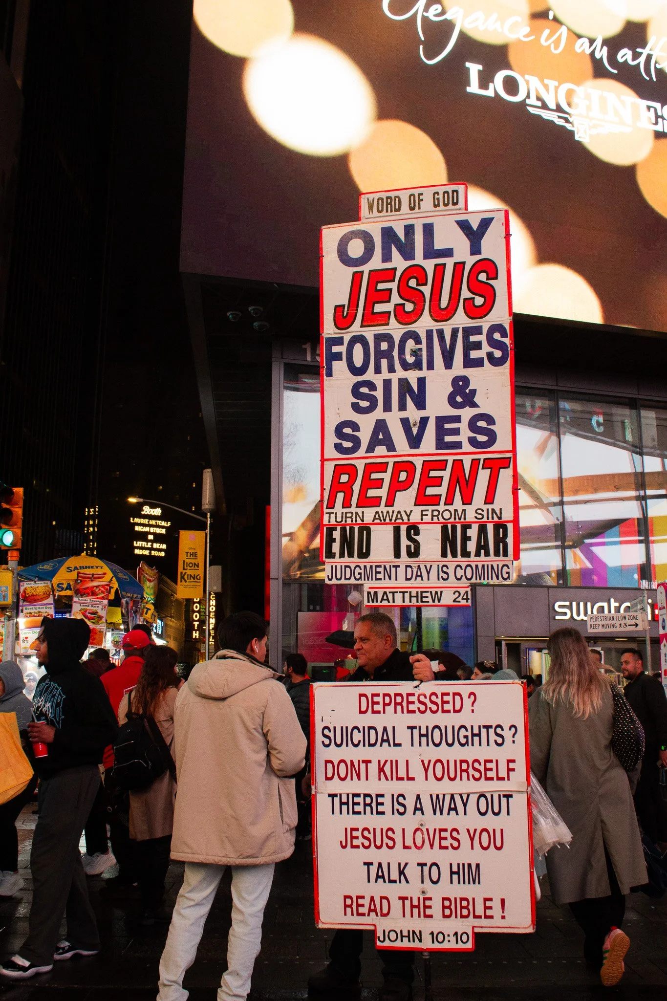 A street scene at night with a crowd of people, two signs with religious messages, and a large digital billboard overhead showing bokeh lights and text.